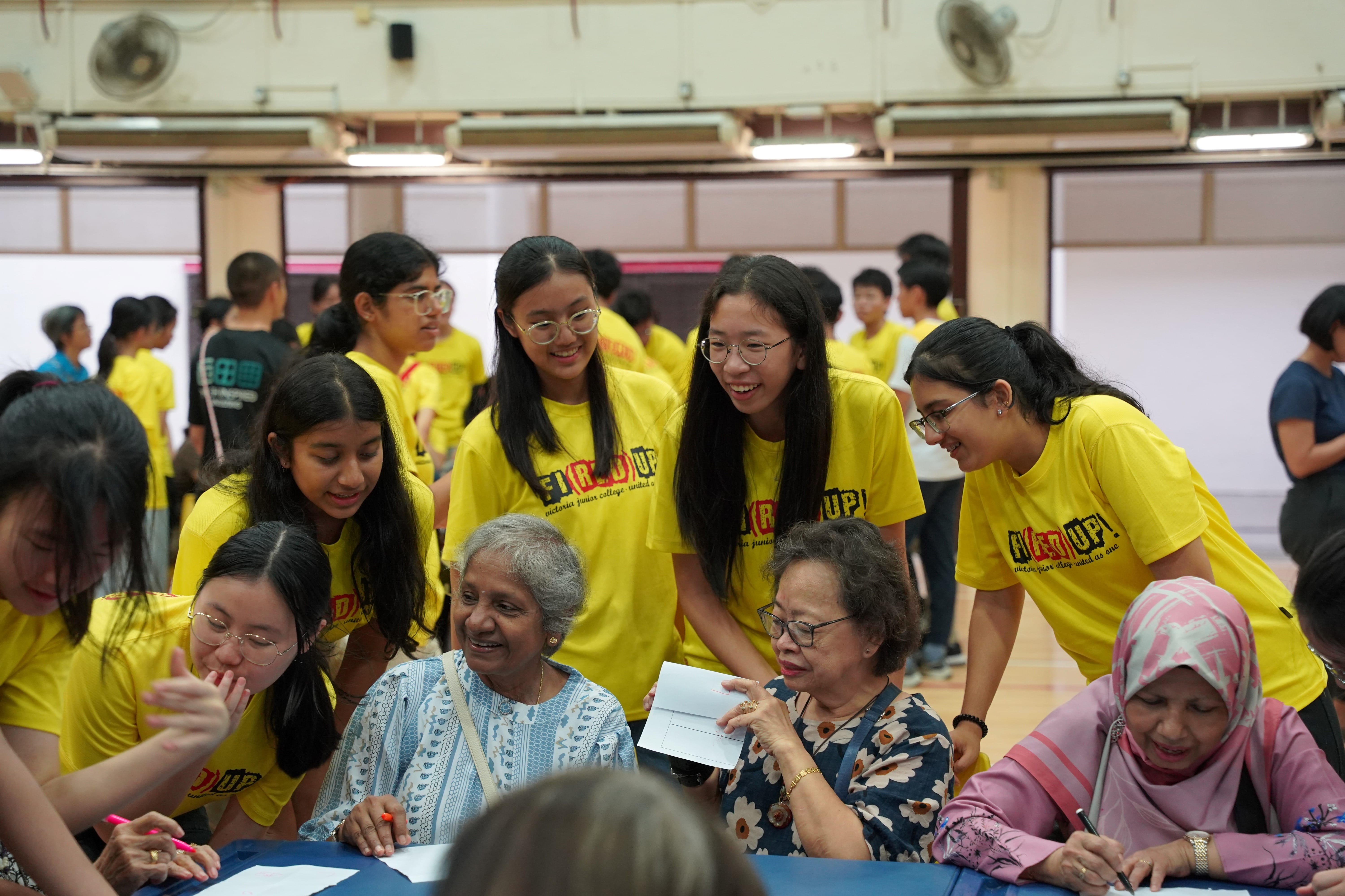 A medium shot shows a heartwarming intergenerational interaction in a brightly lit indoor hall. Three elderly women of different ethnicities are seated at a long blue table in the foreground, engaged in an activity involving writing on papers and envelopes. Surrounding them are several smiling teenage students wearing matching bright yellow t-shirts with the text "FI(RE)D UP!" printed across the front. The students bend down to talk, laugh, and assist the elderly women, creating a joyful, community-oriented atmosphere. In the background, other students in yellow and black t-shirts are scattered across the spacious hall, which is equipped with wall-mounted fans and large windows.
