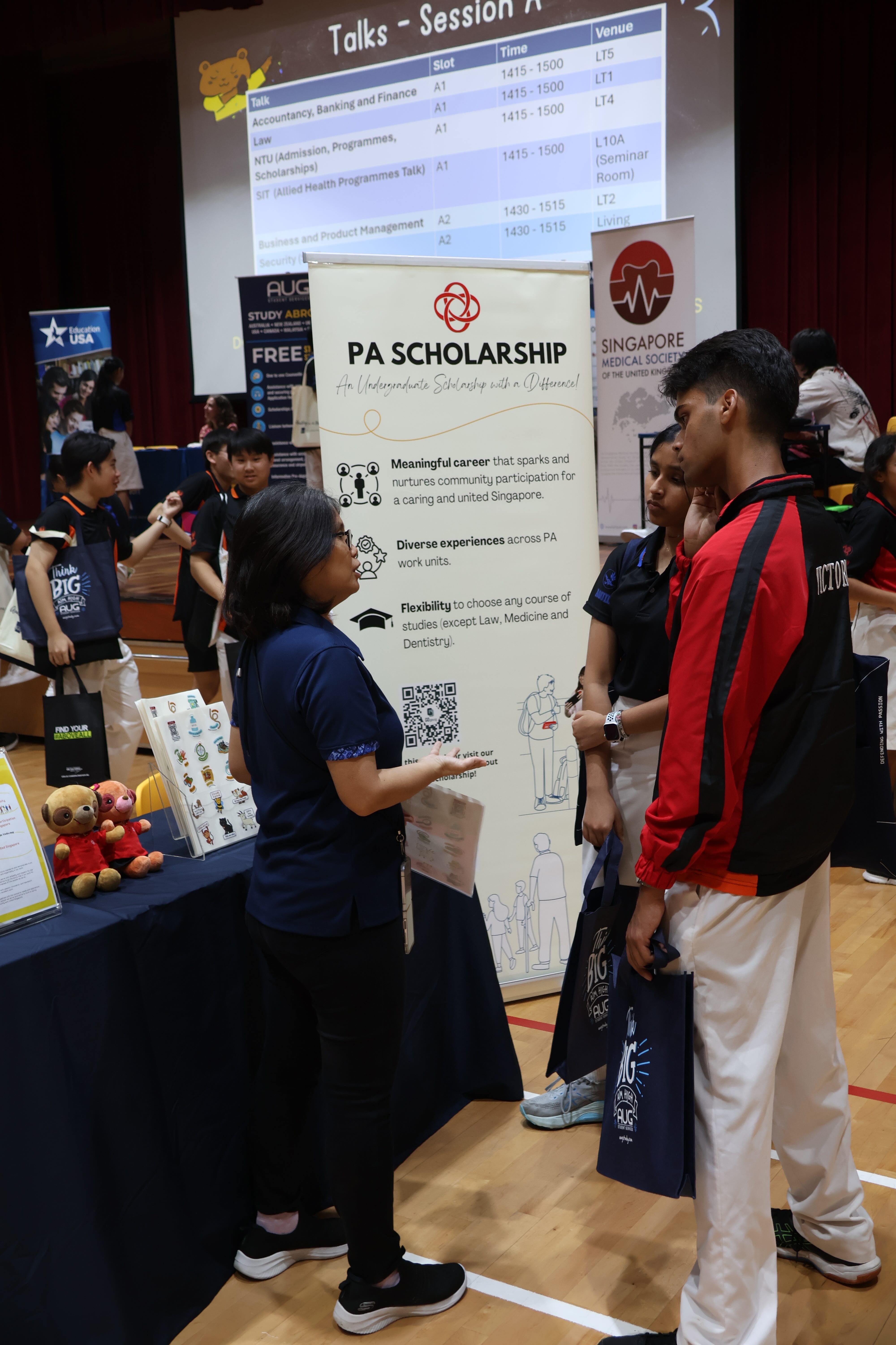 A woman speaks to students at a PA Scholarship booth during an education fair with banners and a projected schedule.
