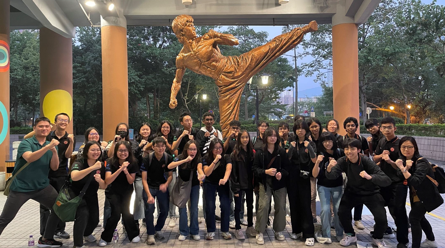 A group of young people in black shirts pose with raised fists before a large bronze martial artist statue outdoors.