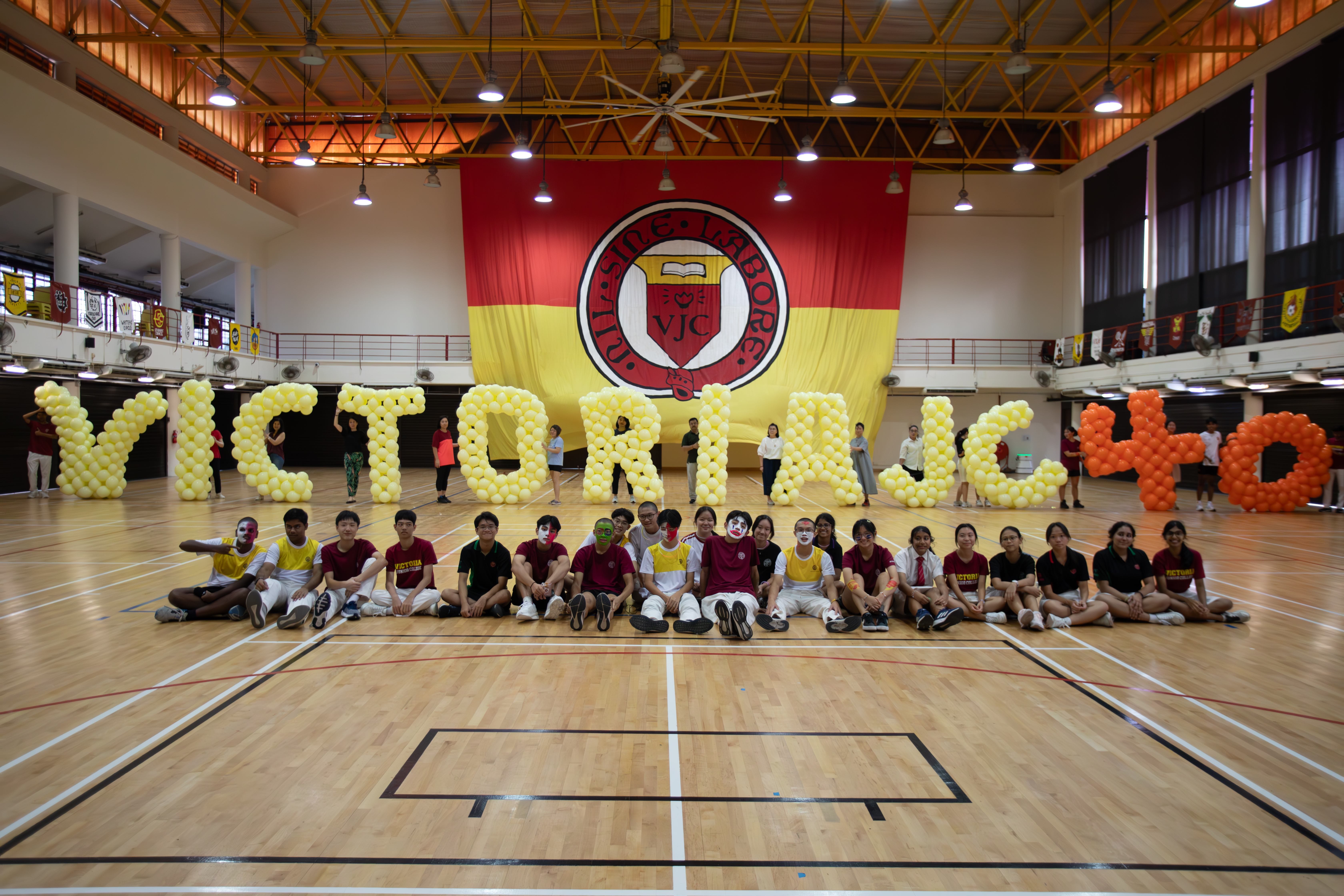 Students sit on a gym floor in front of large balloon letters spelling "Victoria JC" with a VJC school banner behind them.