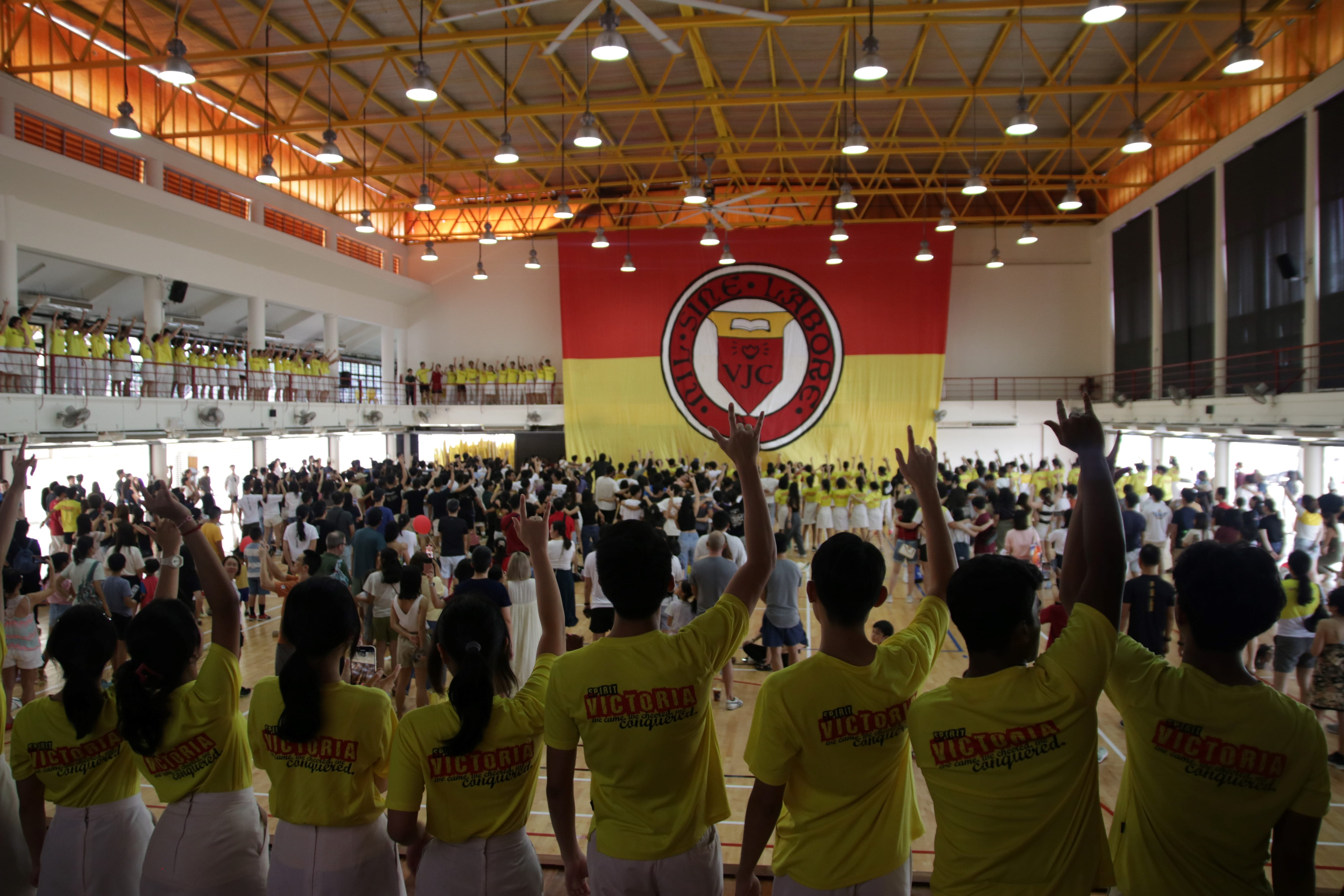 Large crowd in yellow VJC shirts cheering in a gymnasium with a giant red and yellow VJC school banner hanging behind them.