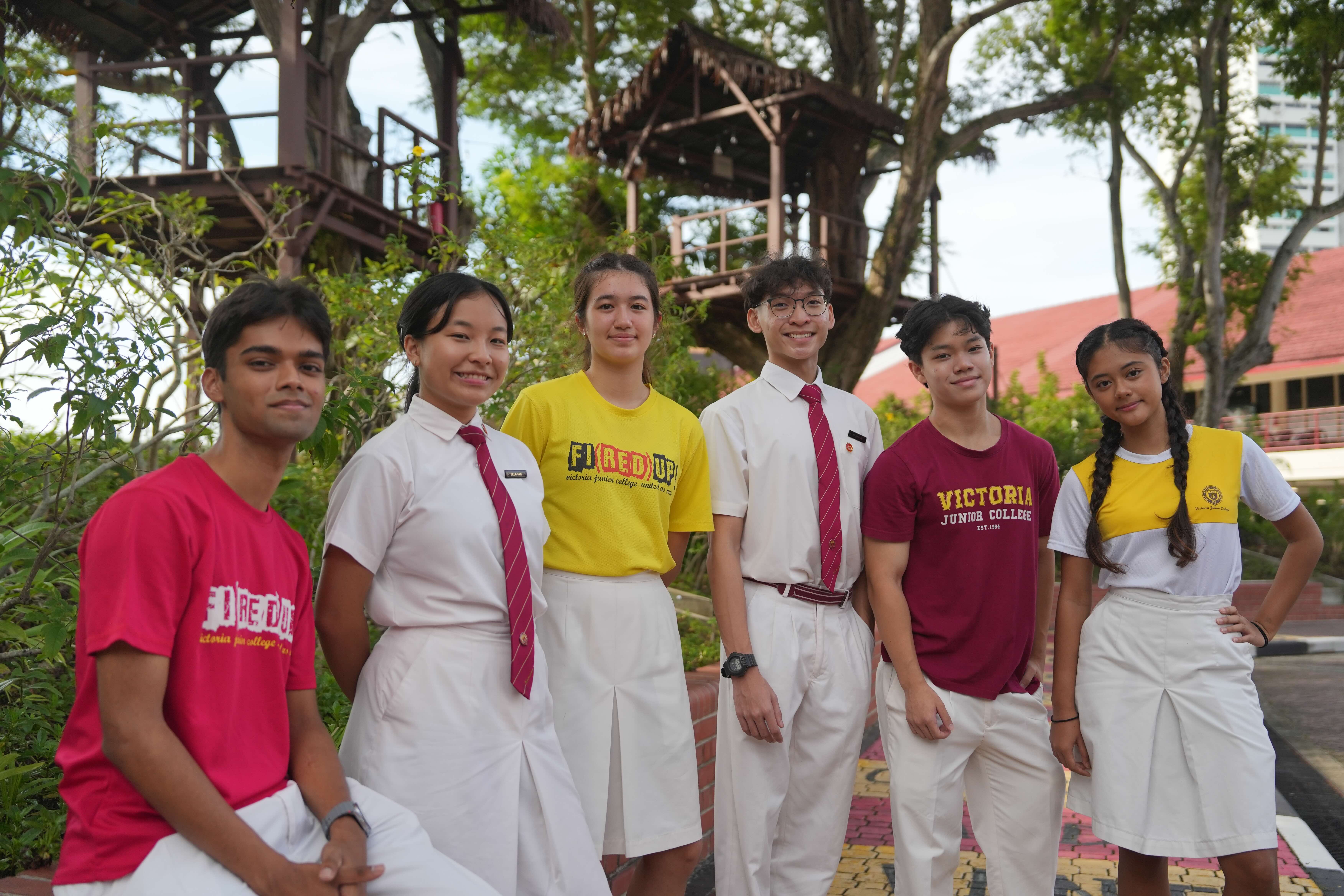 Six Victoria Junior College students in uniforms and school shirts pose outdoors near a treehouse structure.