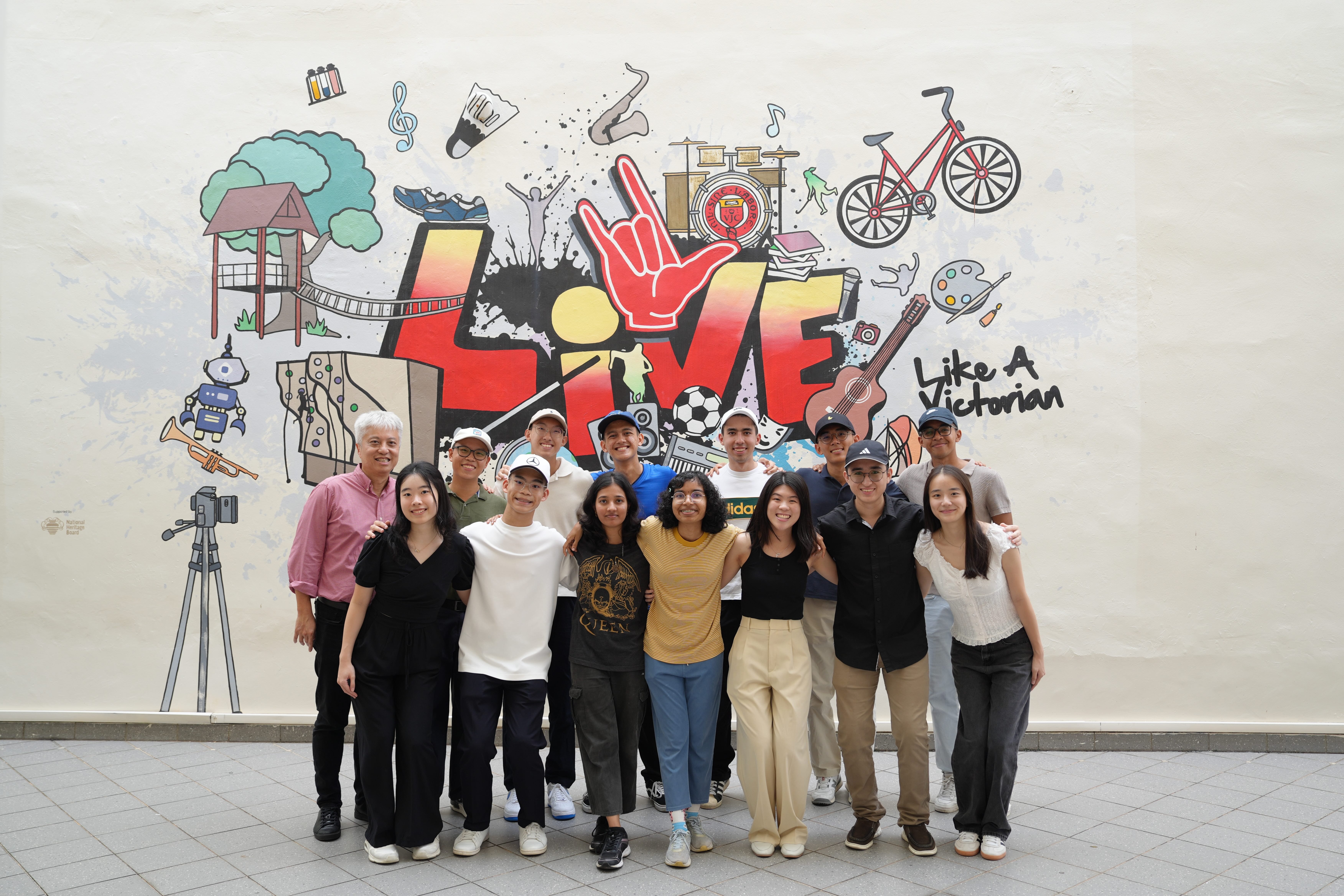 A group of about 13 people posing in front of a colorful mural reading "Live Like A Victorian."