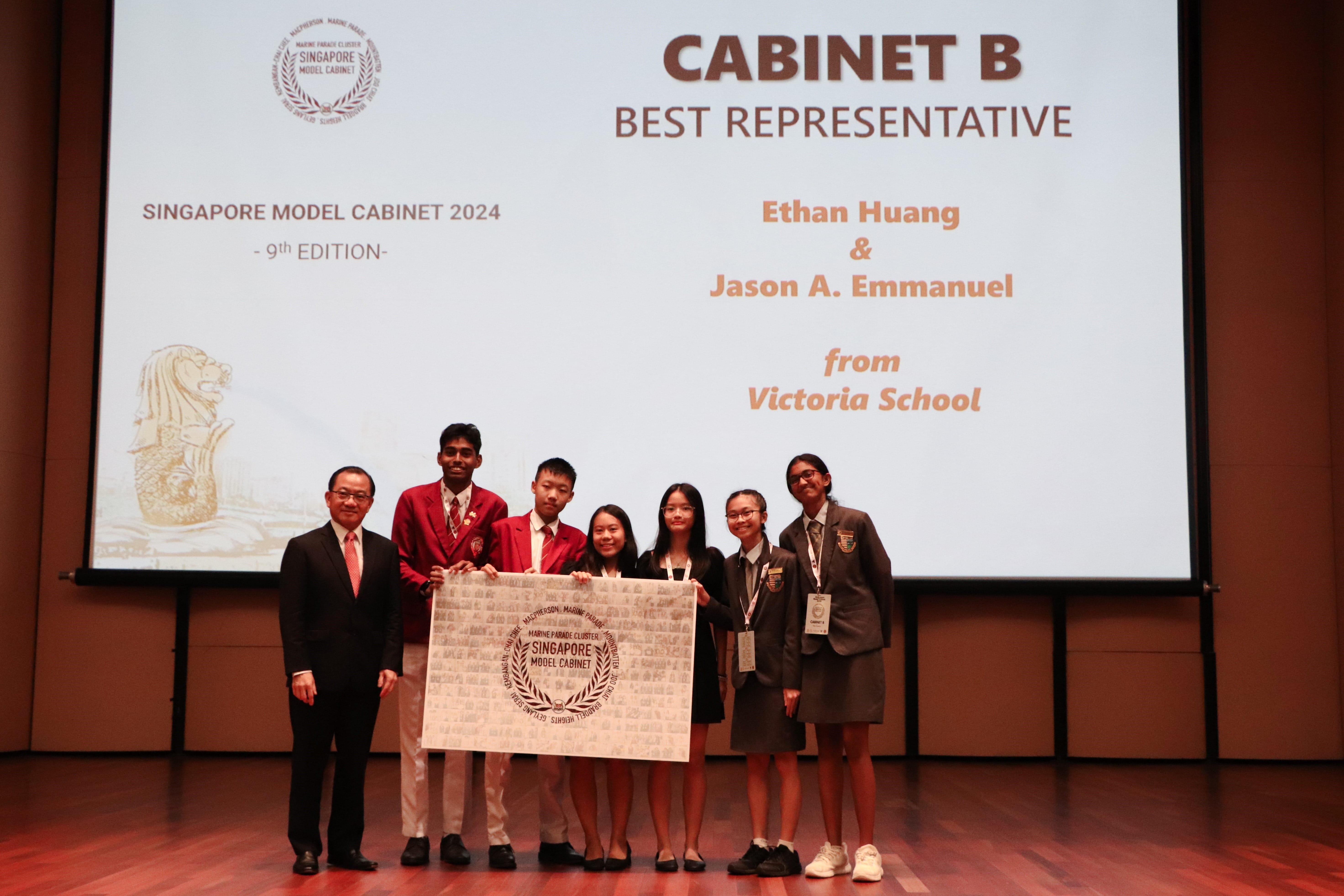 A group of seven people pose for a commemorative photo on a stage in front of a large projection screen during an awards ceremony.