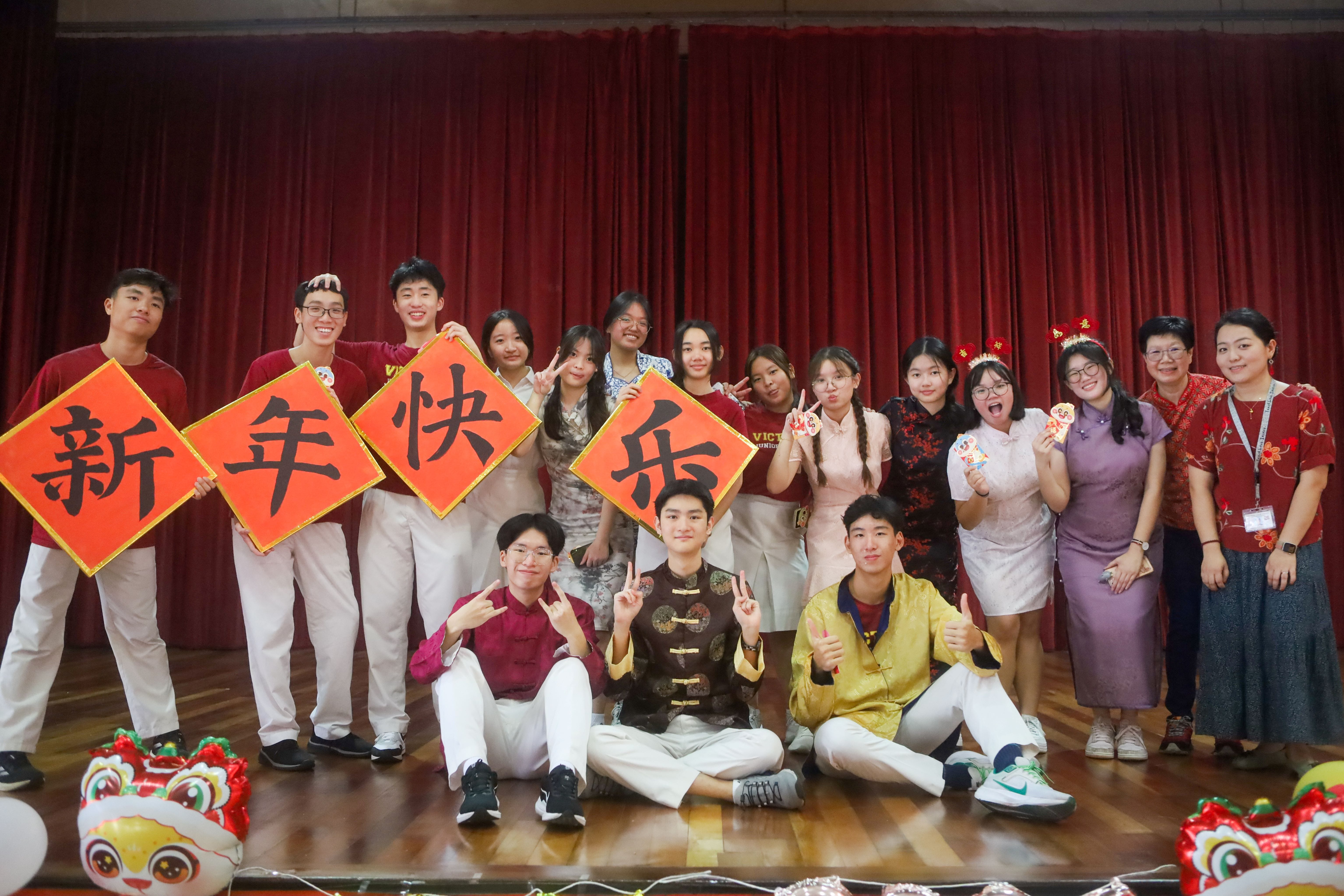 A group of students on stage holding red signs with Chinese characters meaning Happy New Year, dressed in traditional outfits.