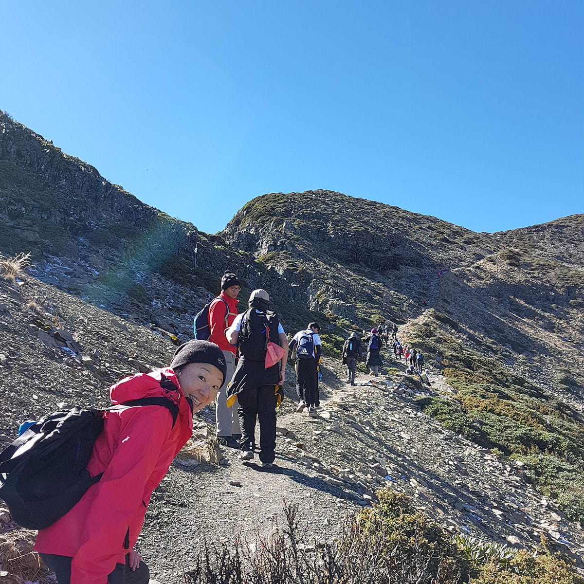 A smiling hiker in a red jacket leads a group climbing a rocky mountain trail under a clear blue sky.