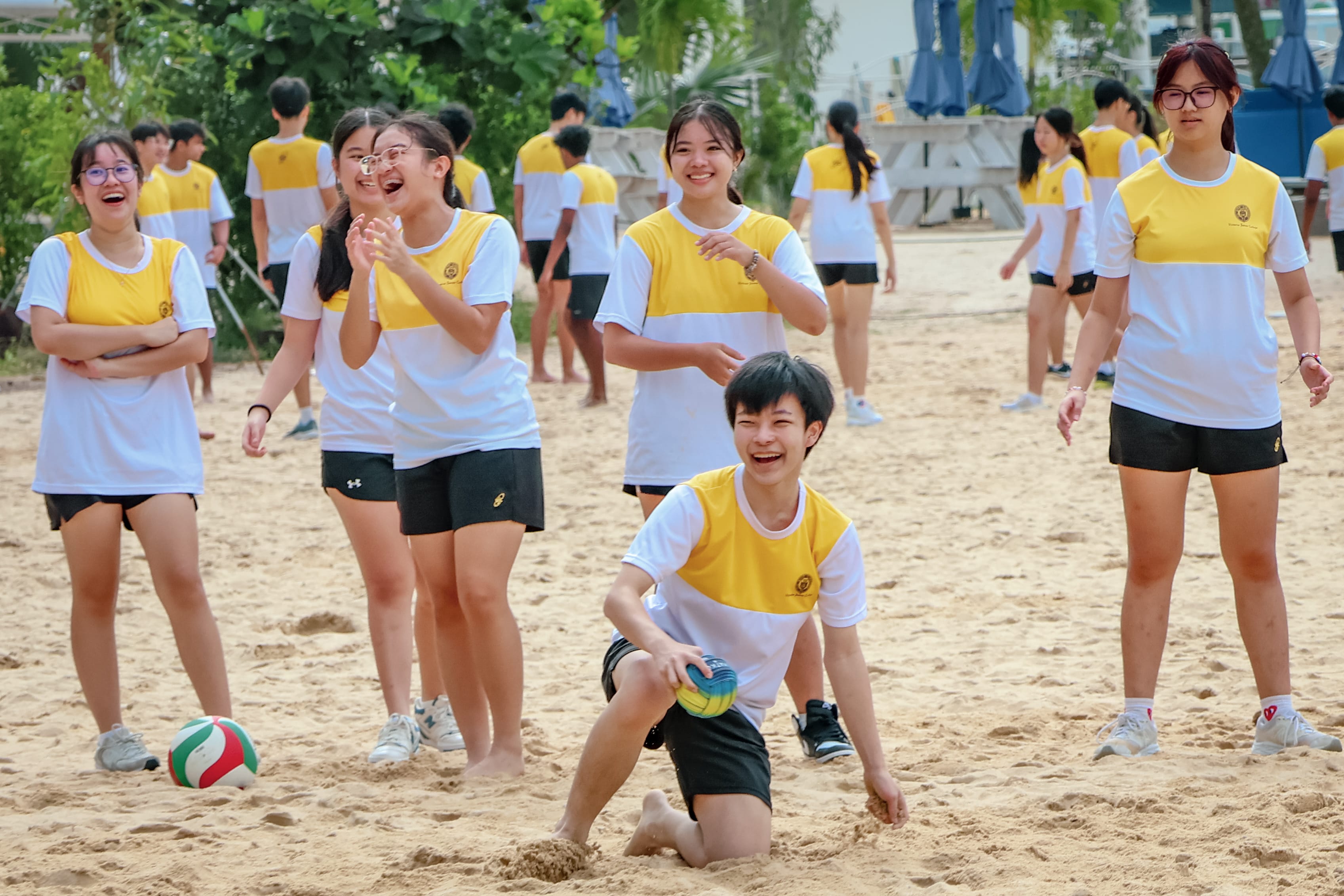 Students in yellow and white uniforms laugh and play volleyball on a sandy outdoor court.