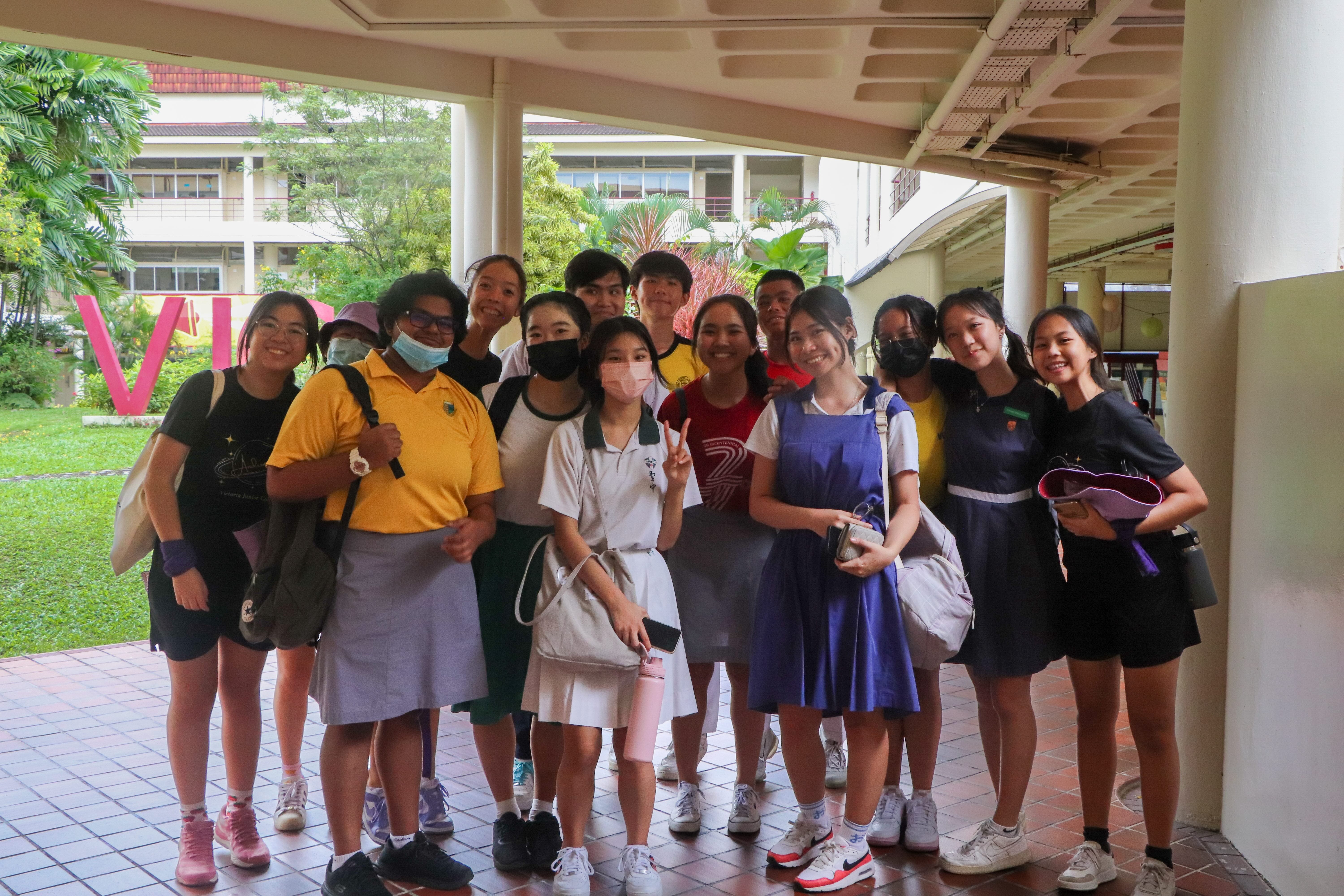 A group of about 13 students in casual and school uniforms pose together under a covered walkway near a school building.