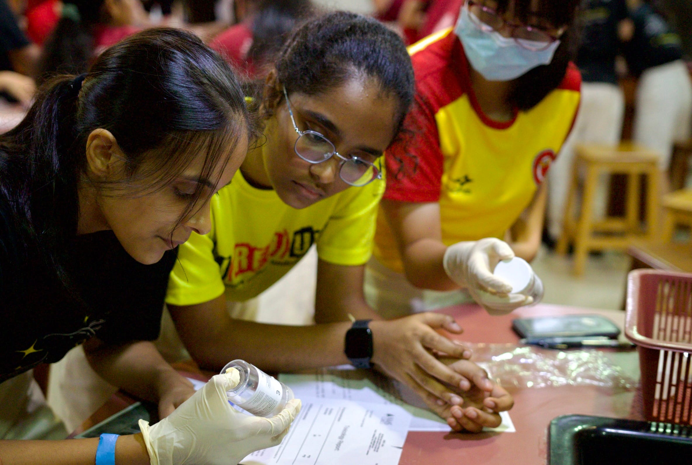 Three girls wearing gloves lean over a table during a hands-on science activity with small white materials.