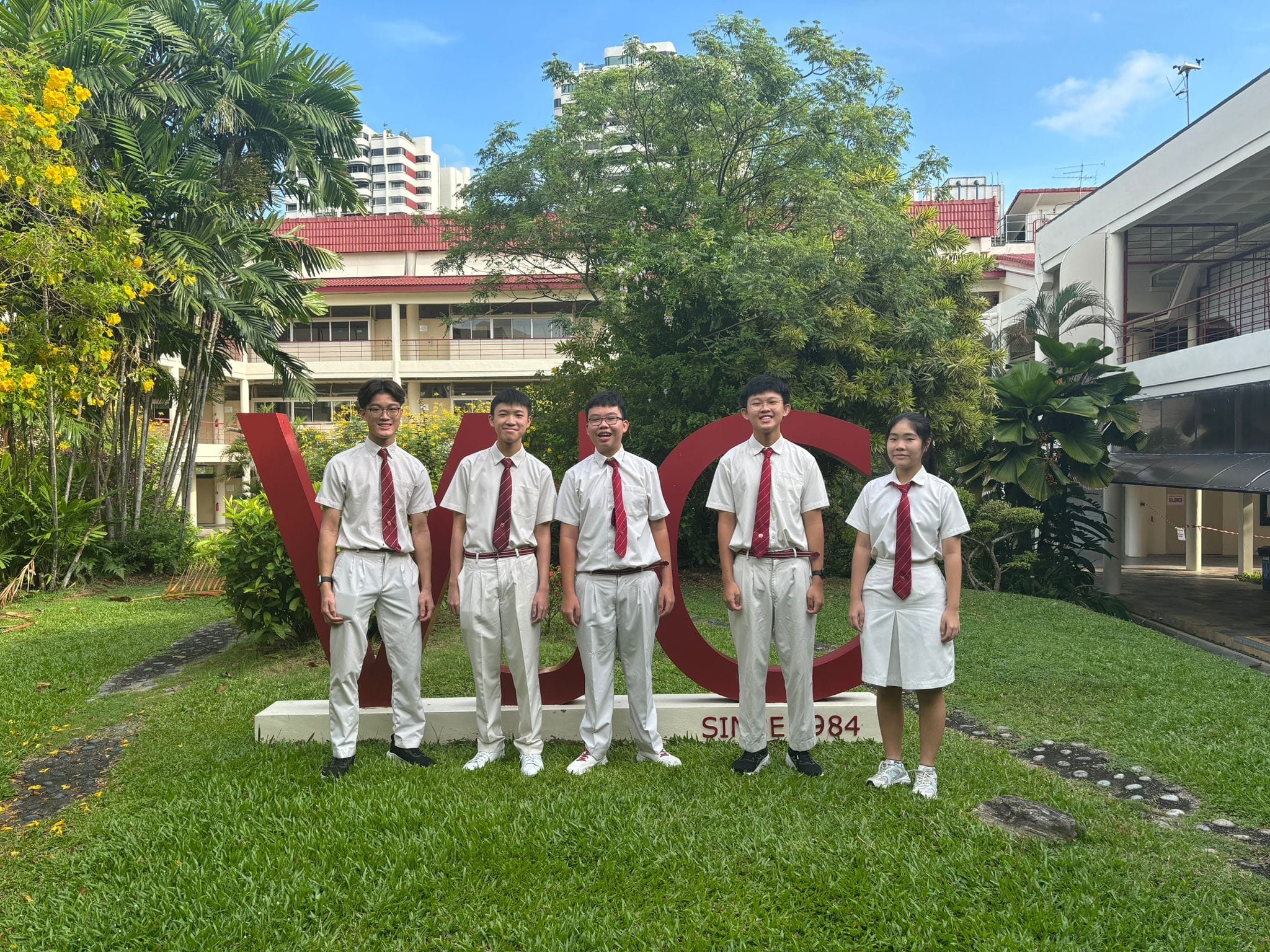 Five students in white uniforms with red ties stand on grass before a large red school sign reading "Since 1984."