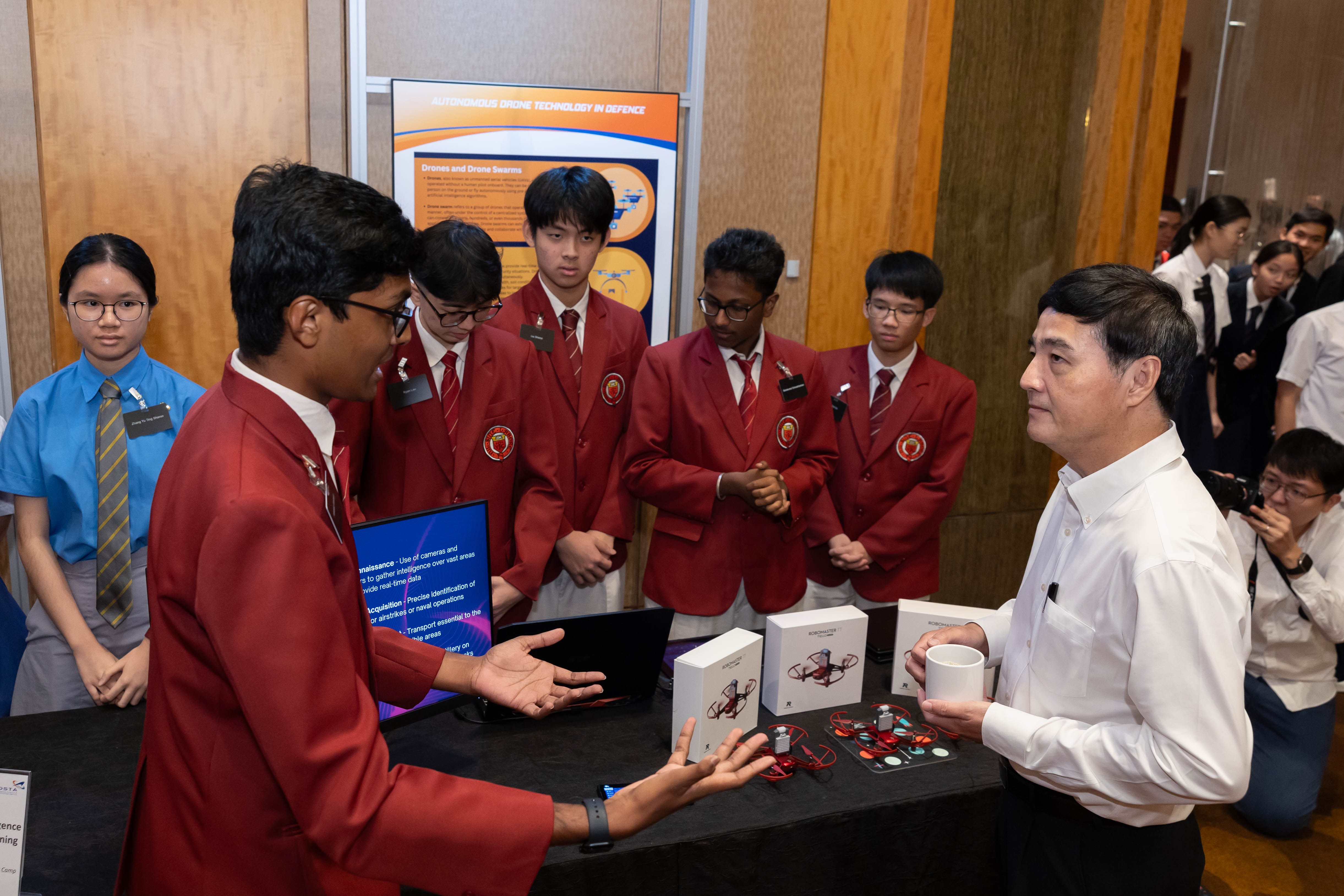 Students in red blazers present a drone technology exhibit to an adult man at an indoor science fair.