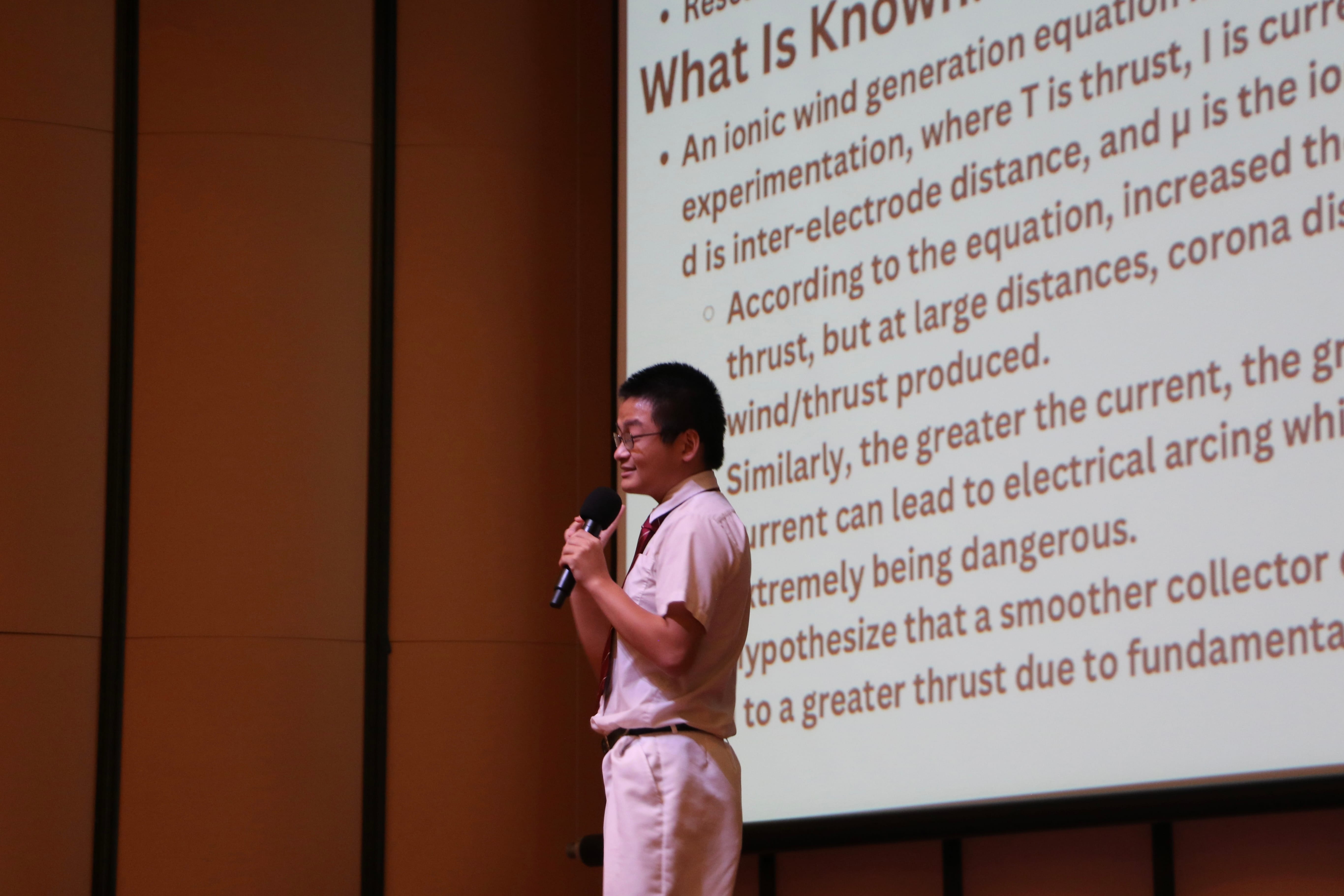 A young man holding a microphone presents in front of a large projected slide about ionic wind generation.