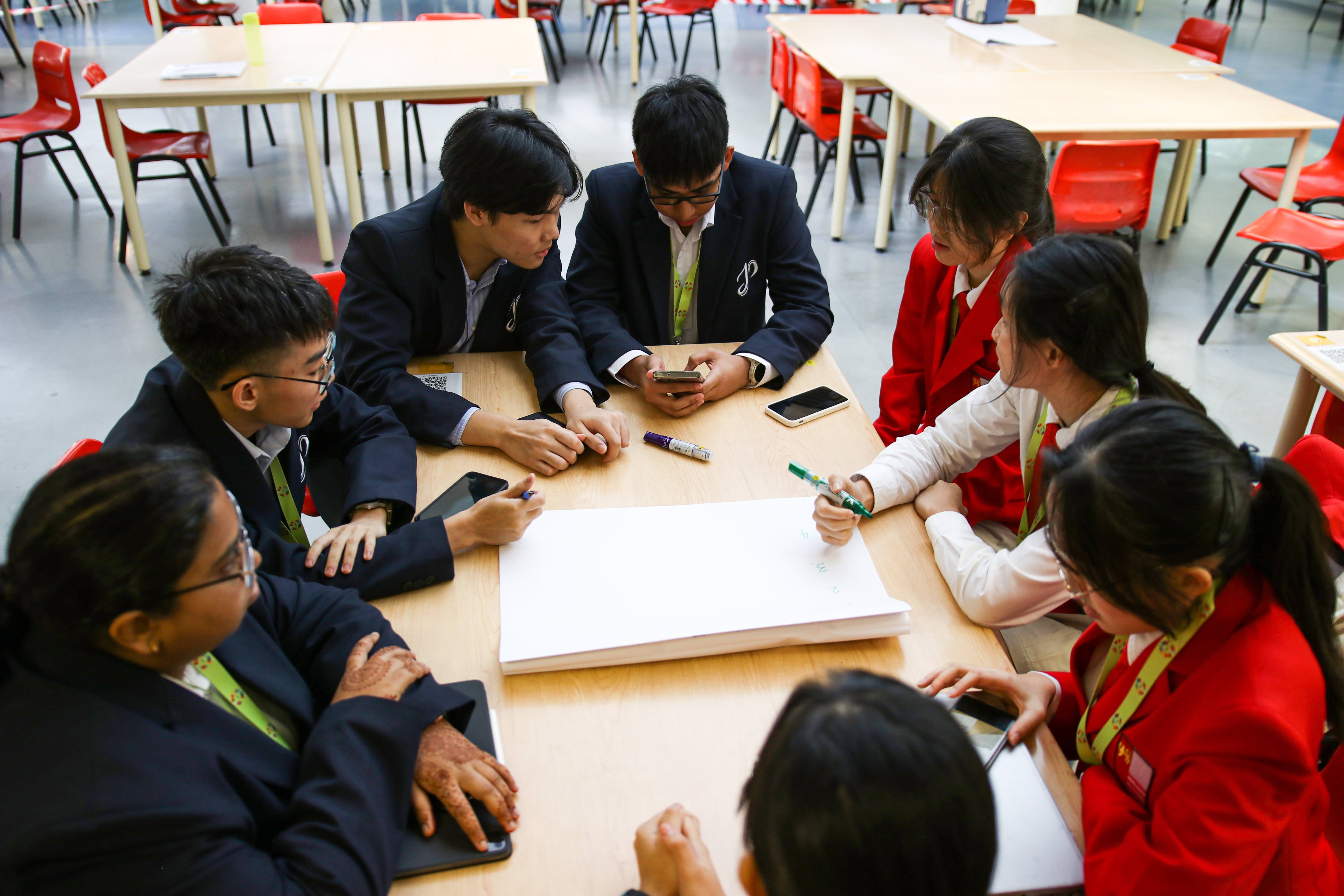 Seven uniformed students in navy and red blazers sit around a table collaborating, with papers and phones visible.