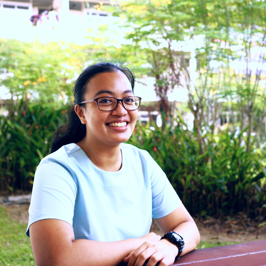 A smiling young woman with glasses and a light blue shirt sits outdoors with green plants in the background.