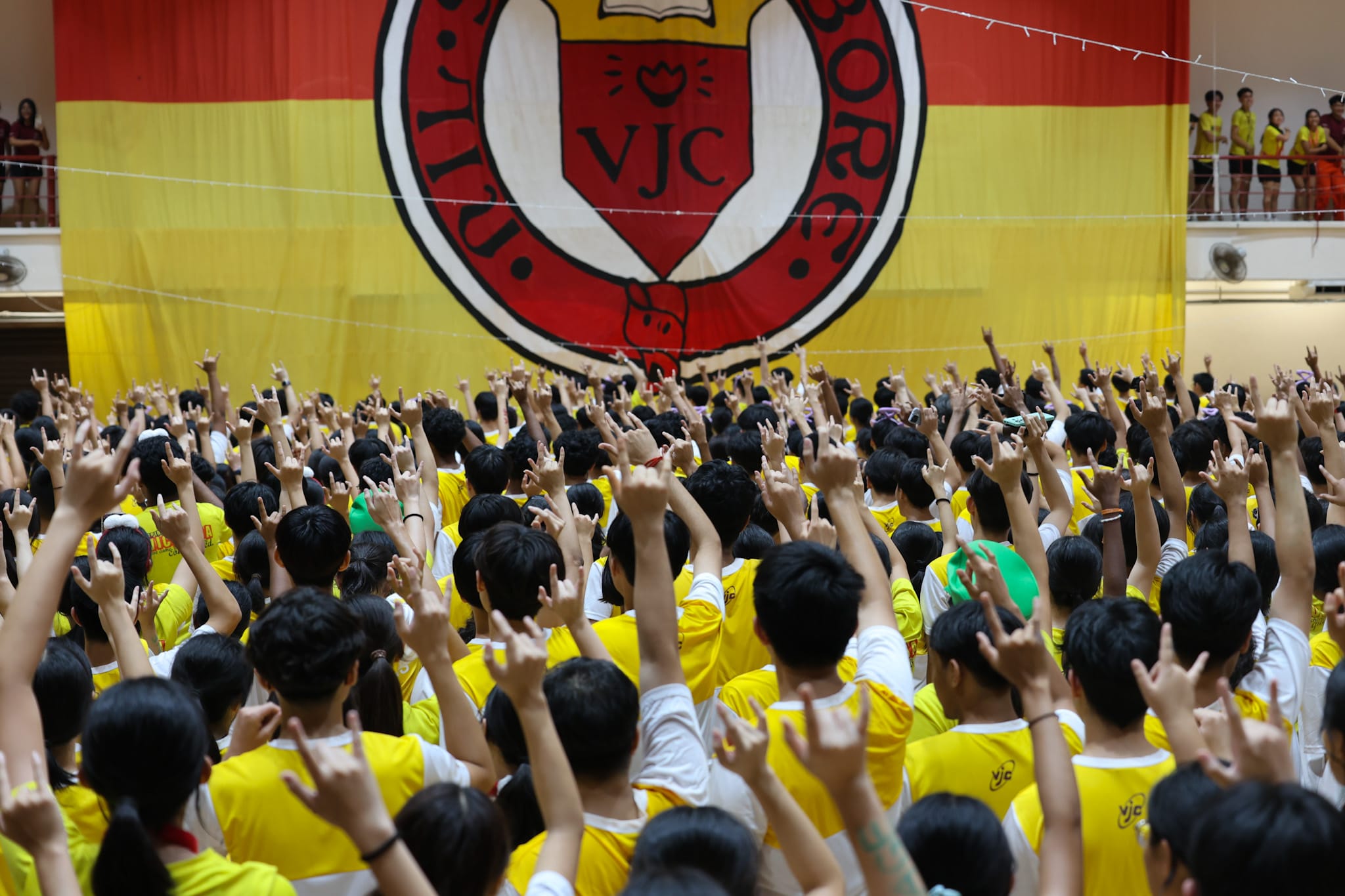 A large crowd in yellow shirts raises hands before a big red and yellow VJC school banner.
