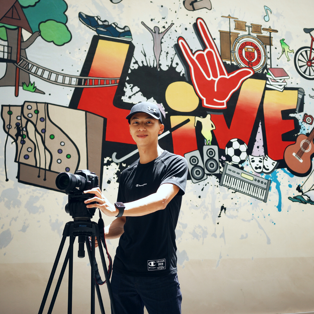 Young man in a black shirt and cap holds a Canon camera on a tripod in front of a colorful mural.