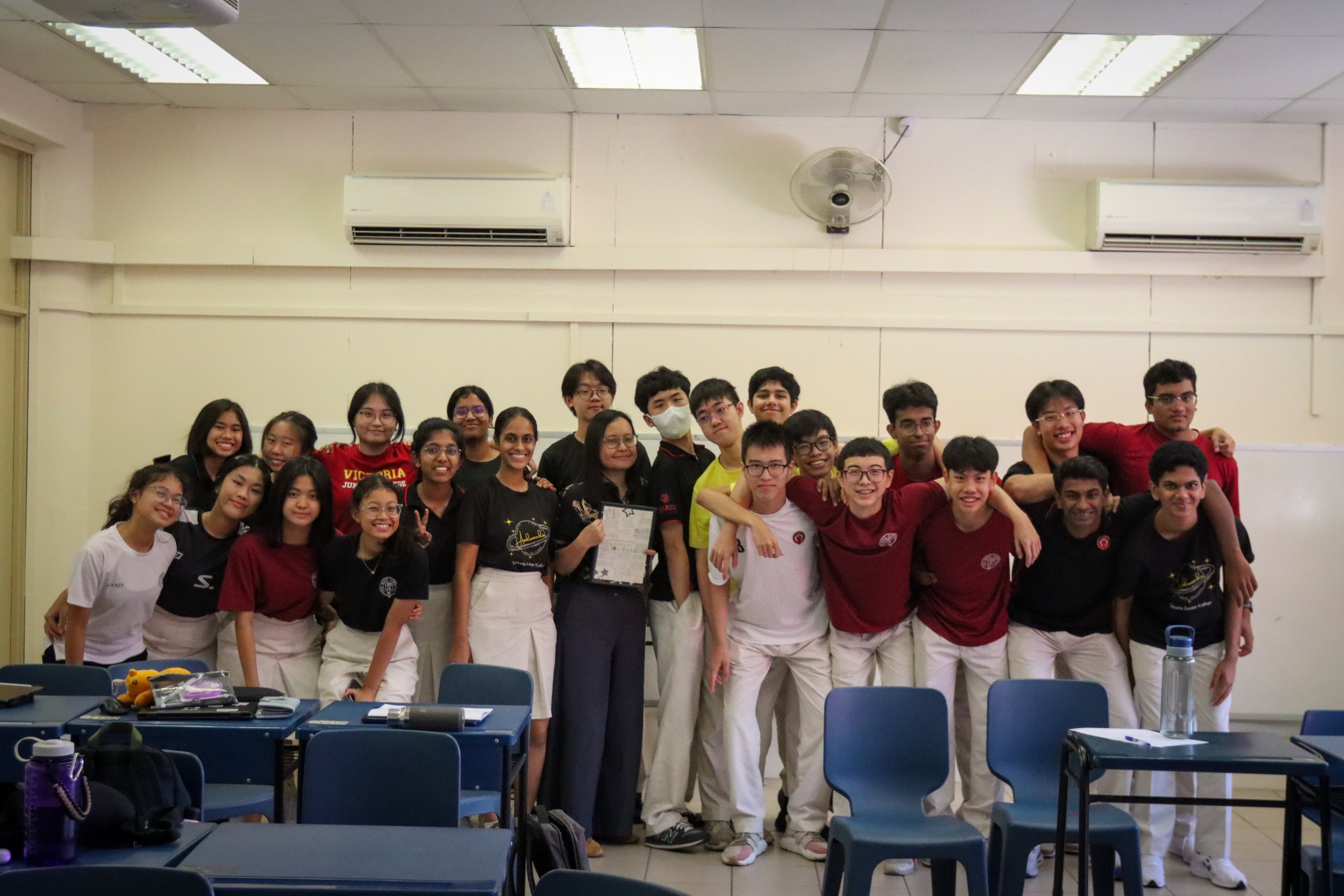 A large group of students posing together in a classroom, wearing black, red, and white outfits.