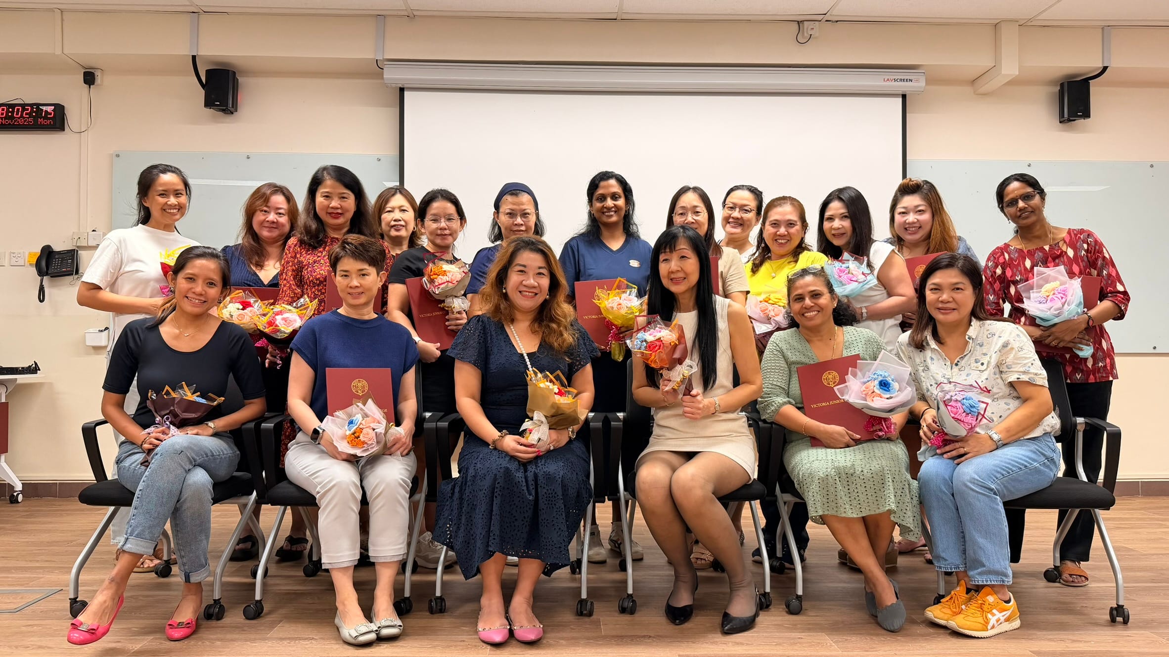 A group of about 20 women posing in a classroom, holding colorful flower bouquets and red certificate folders.