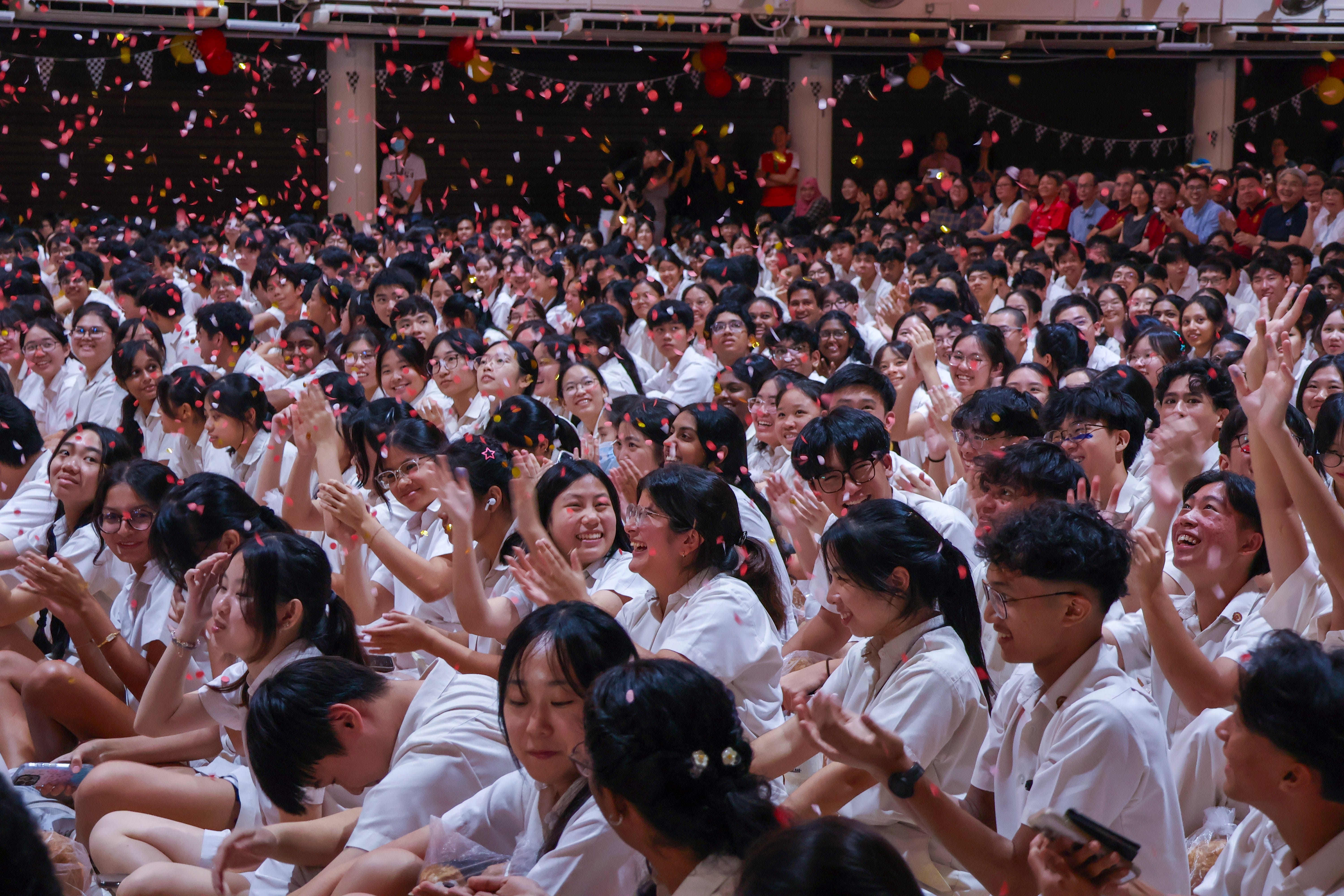 An expansive, wide shot captures a massive, packed crowd of students seated on the floor of a large indoor hall or auditorium, celebrating during an event. The students, primarily of East Asian ethnicity, are all wearing matching white uniform shirts. Red and pink confetti rains down from above across the entire scene, adding a dynamic sense of celebration. Most of the students are smiling, laughing, or clapping, with a few looking up or raised-hands signaling excitement. In the background, tiered seating is filled with a large audience of adults and onlookers watching the event. The atmosphere is vibrant, high-energy, and joyous.