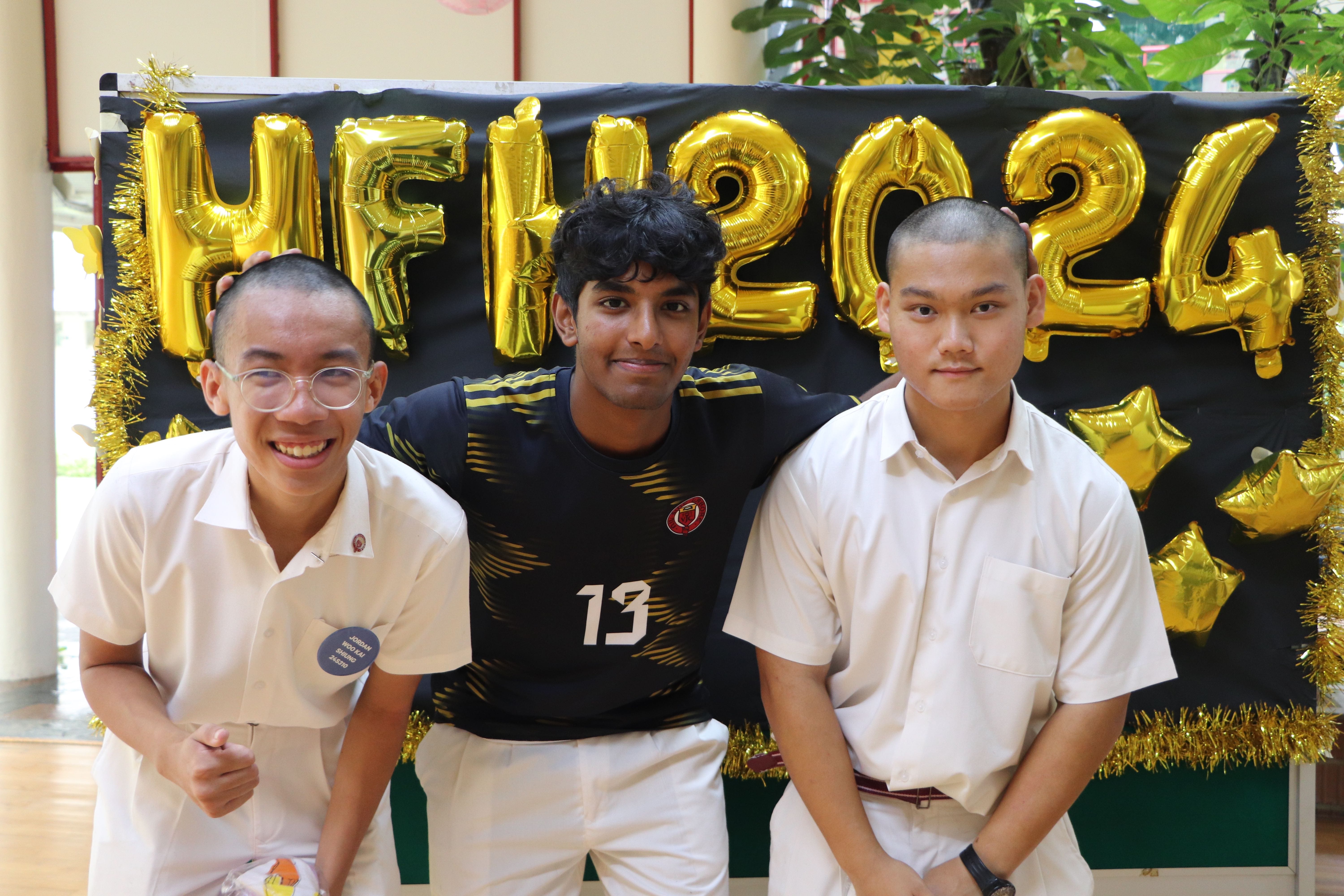 Three teenage boys posing in front of a black backdrop with gold balloons spelling HFH2024.
