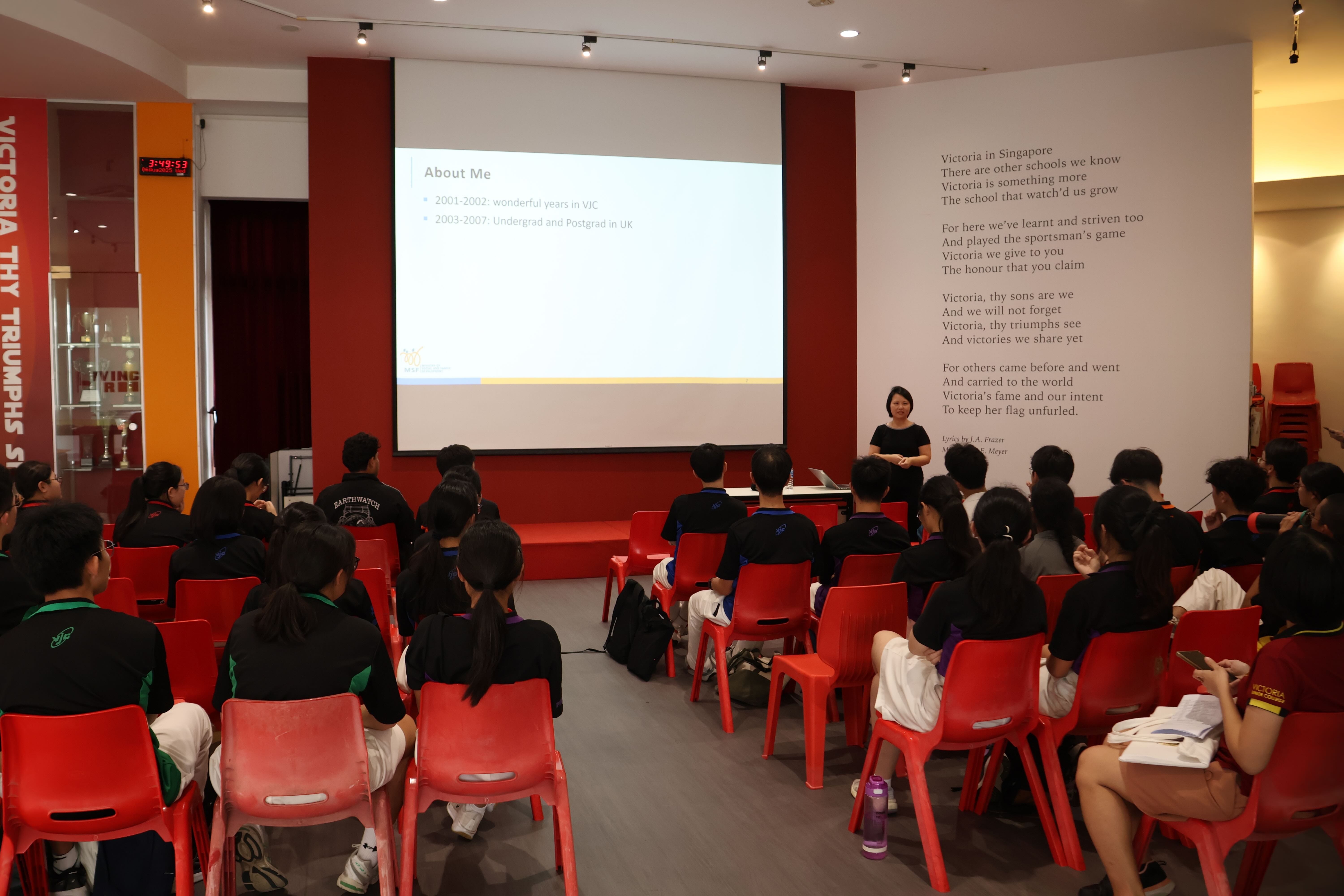 A woman presents an "About Me" slideshow to students seated on red chairs in a school hall with a poem on the wall.