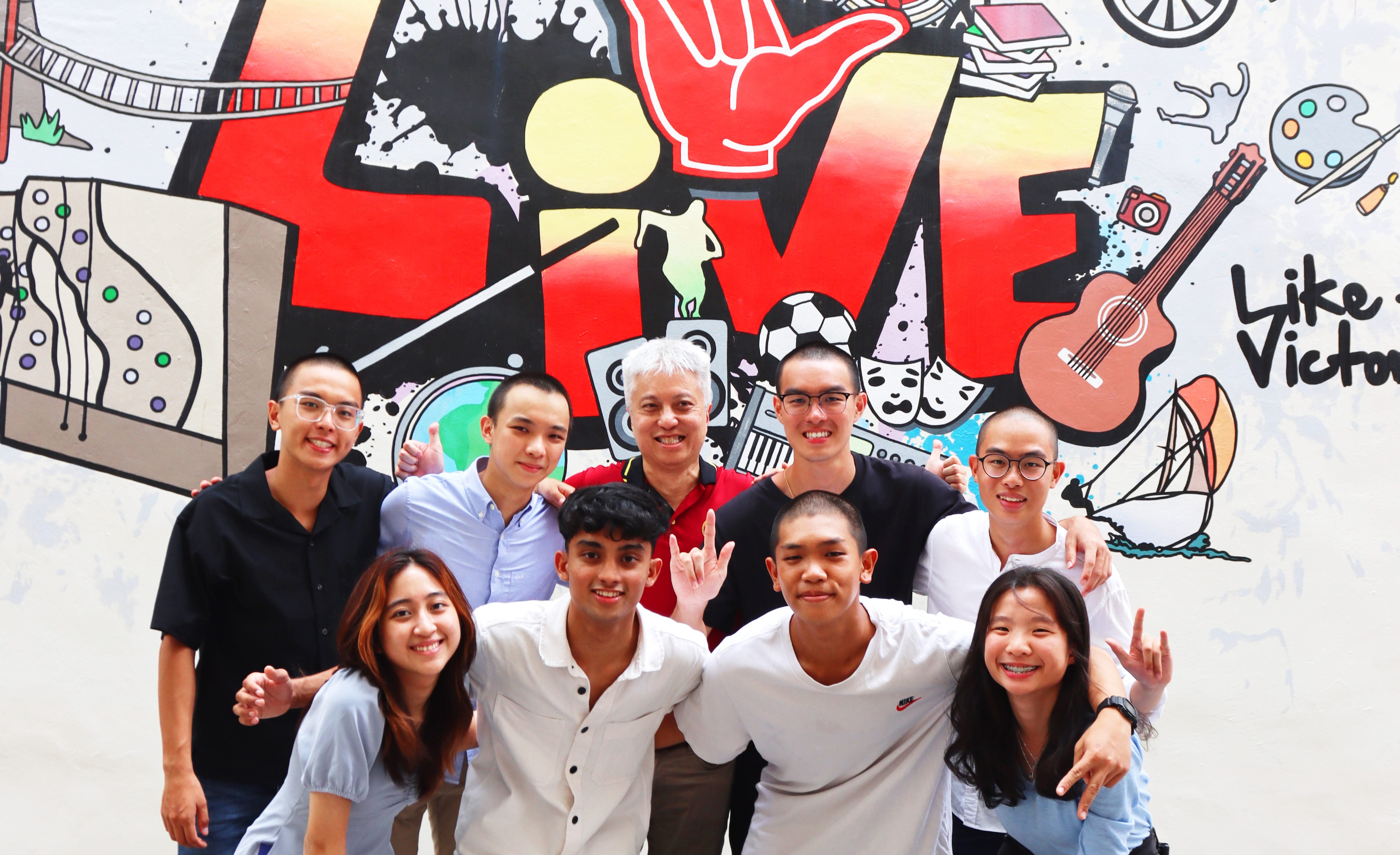 Eight smiling people pose together in front of a colorful mural that reads LIVE with drawings of a guitar and soccer ball.