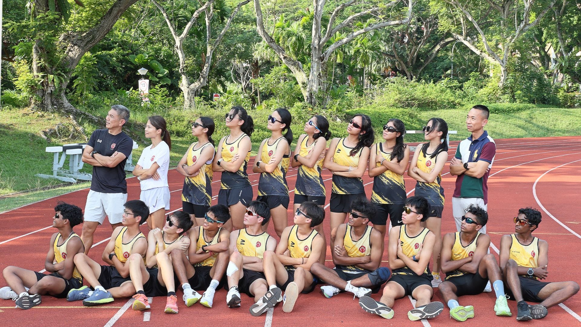 A youth athletics team in yellow and black uniforms poses on a red track with two coaches, surrounded by trees.