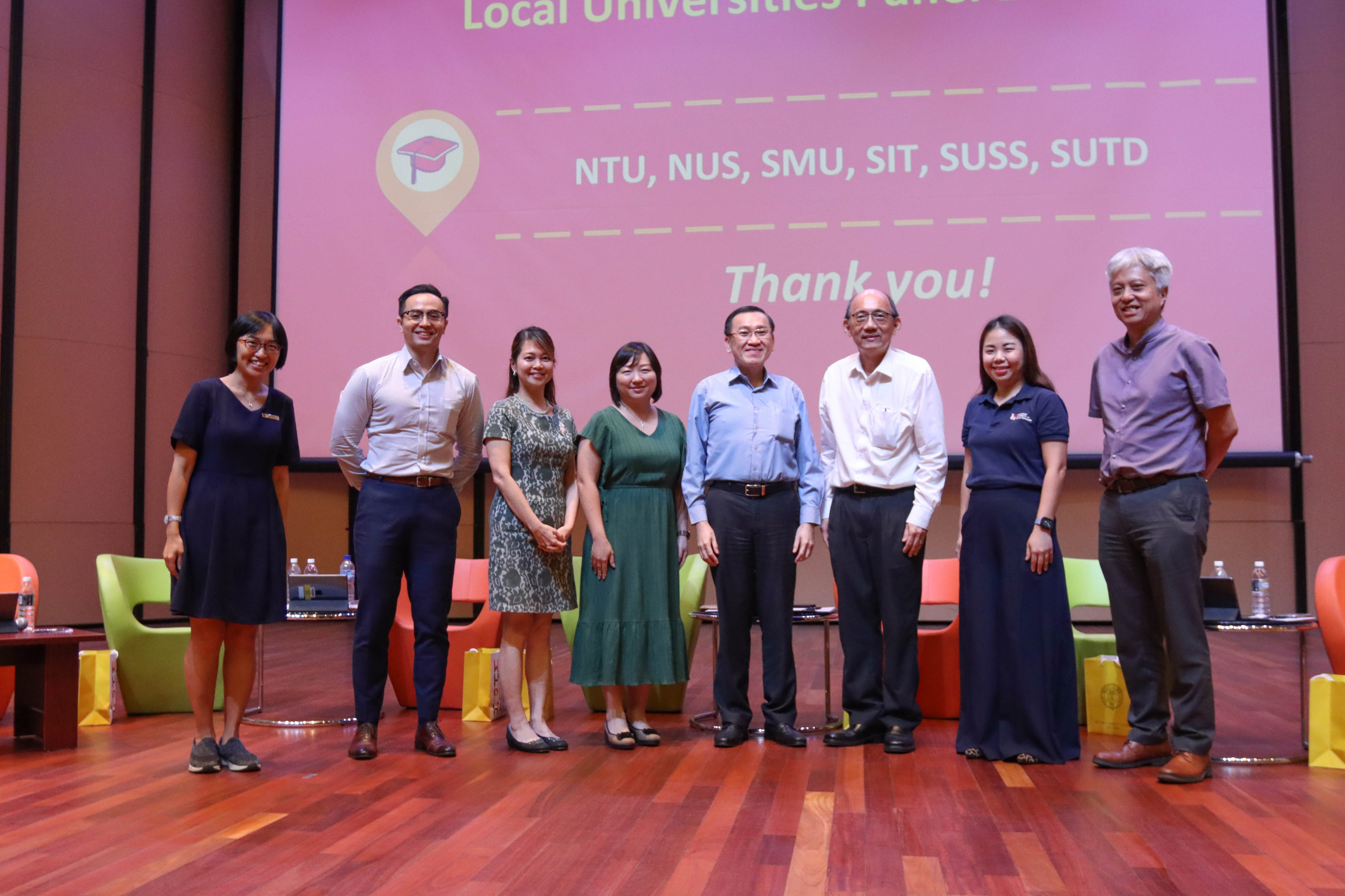 Seven people stand on a stage in front of a pink screen listing NTU, NUS, SMU, SIT, SUSS, SUTD with "Thank you!"