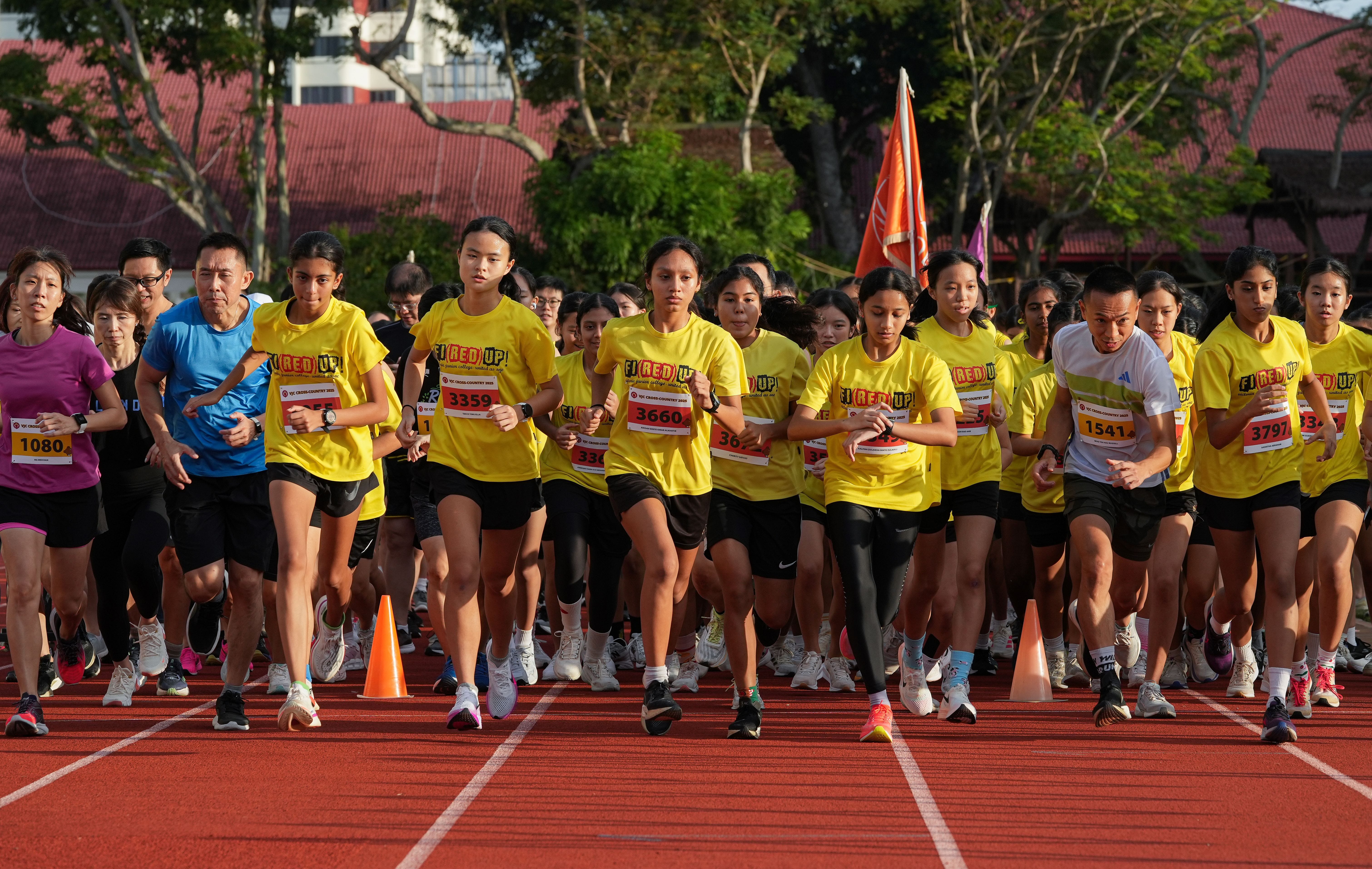 A large group of runners in yellow shirts with race bibs sprint from the start line on a red track.