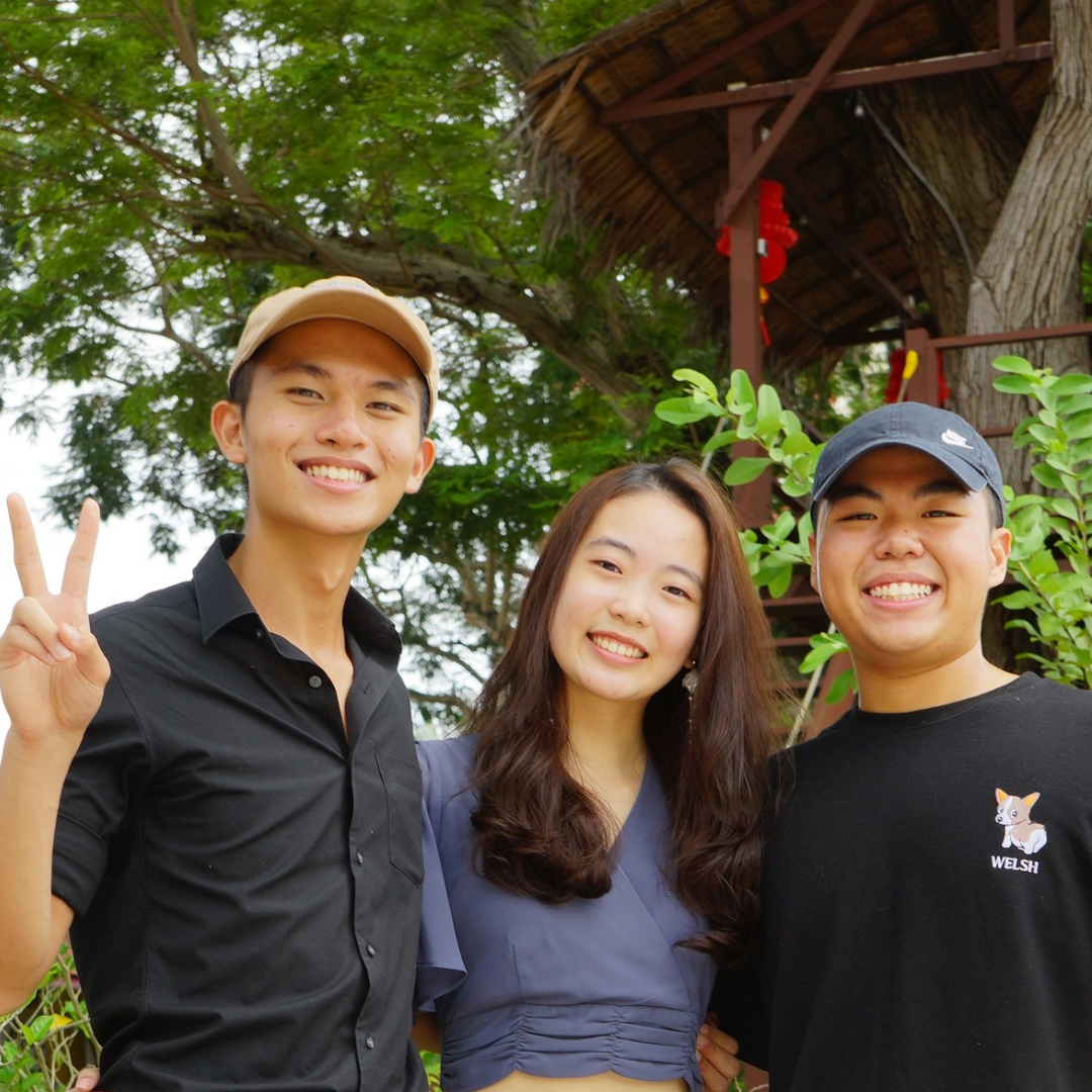 Three smiling young people pose outdoors near a thatched treehouse, with one making a peace sign.