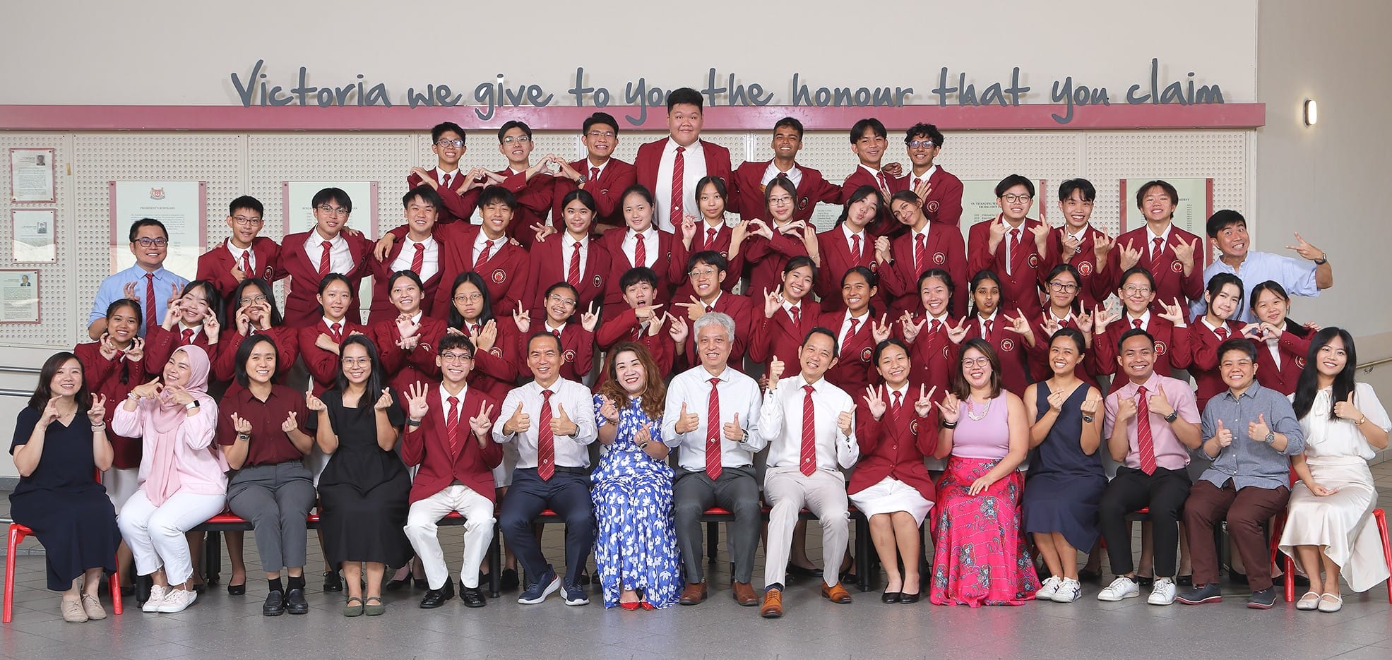 Large group of students and teachers in red blazers posing indoors under a Victoria School motto on the wall.