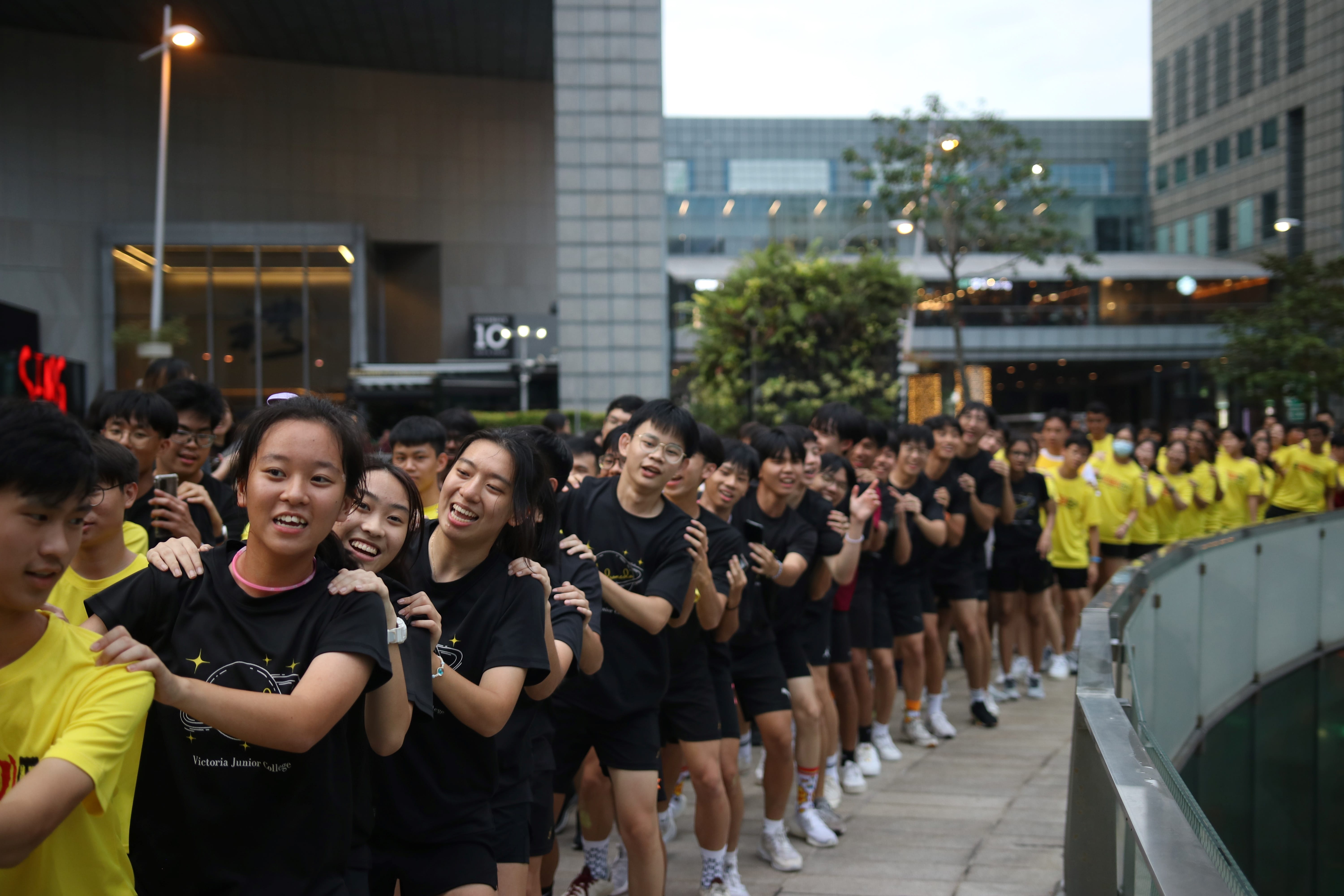 A large group of smiling students in black and yellow shirts lined up outdoors near modern buildings.