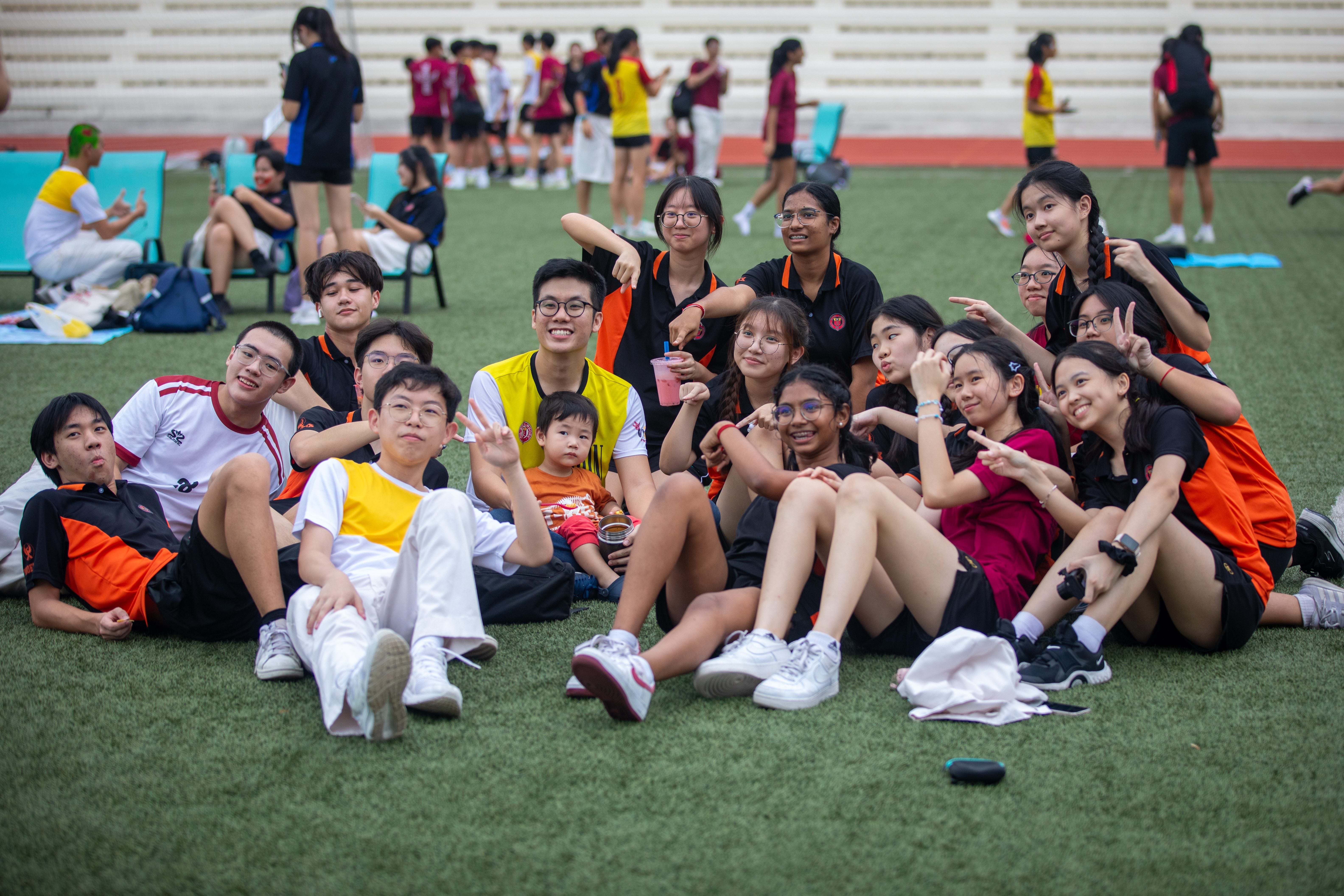 A large group of smiling students in black and orange uniforms sit together on a green field at a sports event.