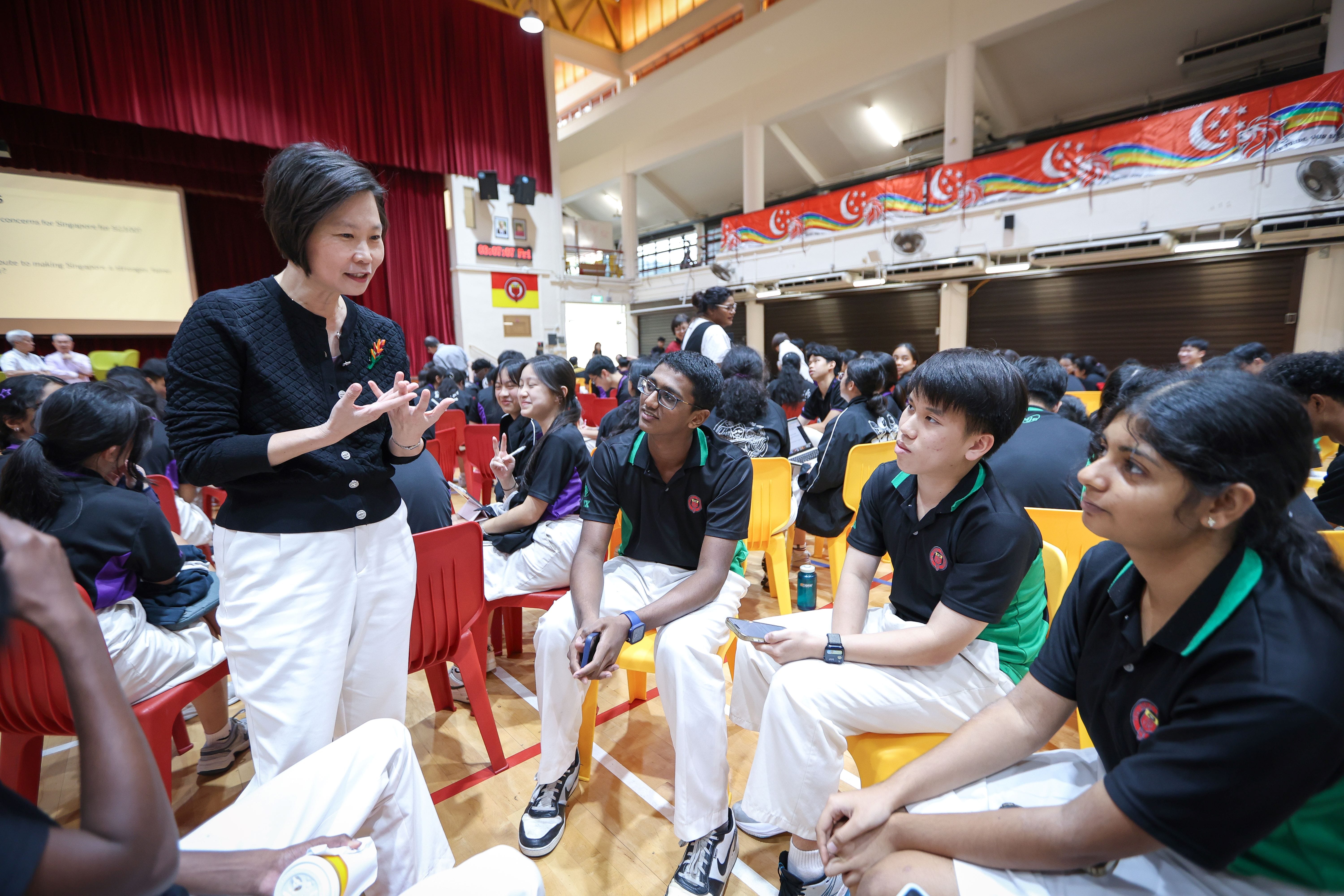 A woman in a black top and white pants speaks to uniformed students seated on colorful chairs in a school hall.