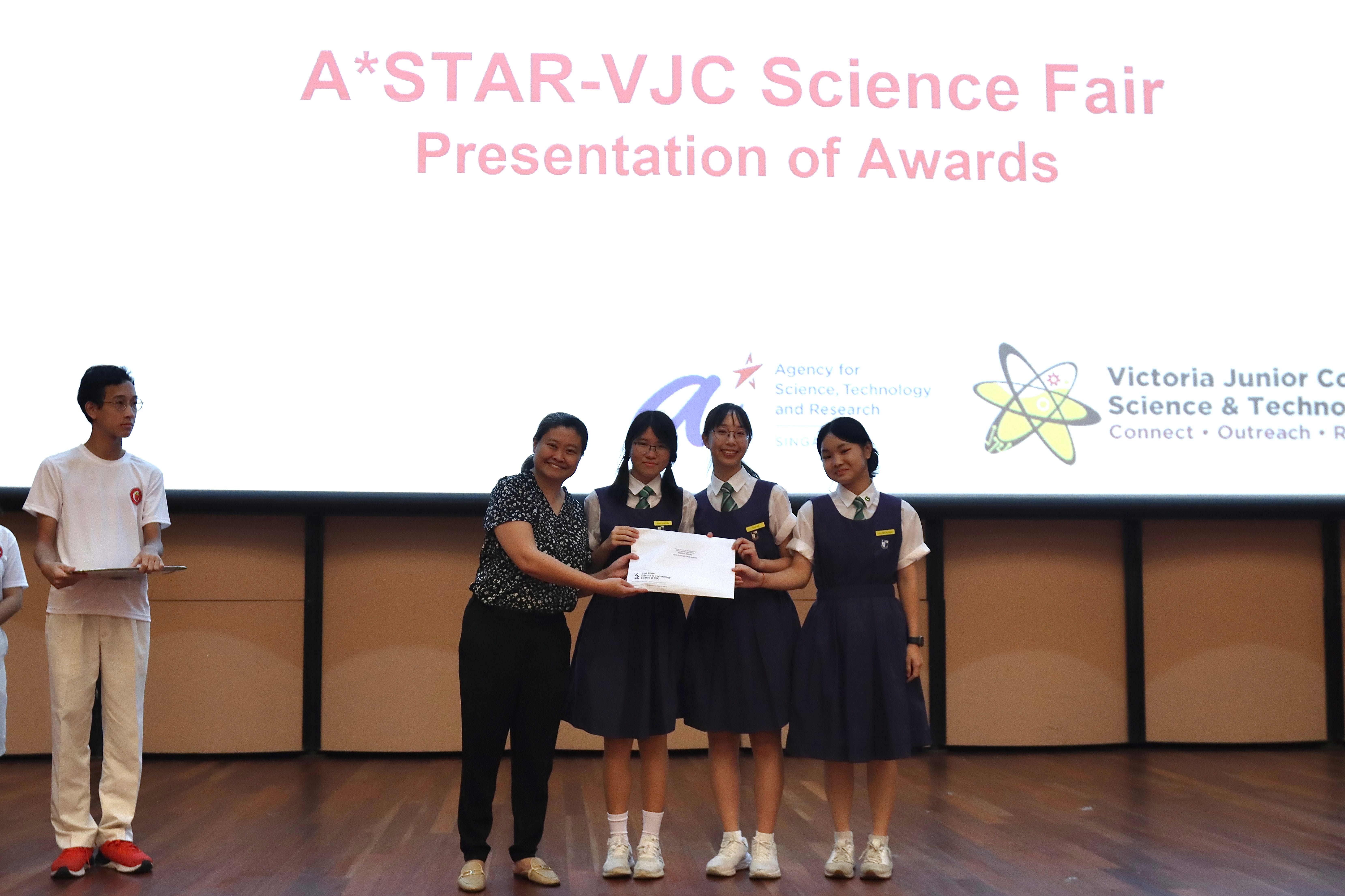An adult presents a certificate to three uniformed students on stage at the A*STAR-VJC Science Fair awards ceremony.