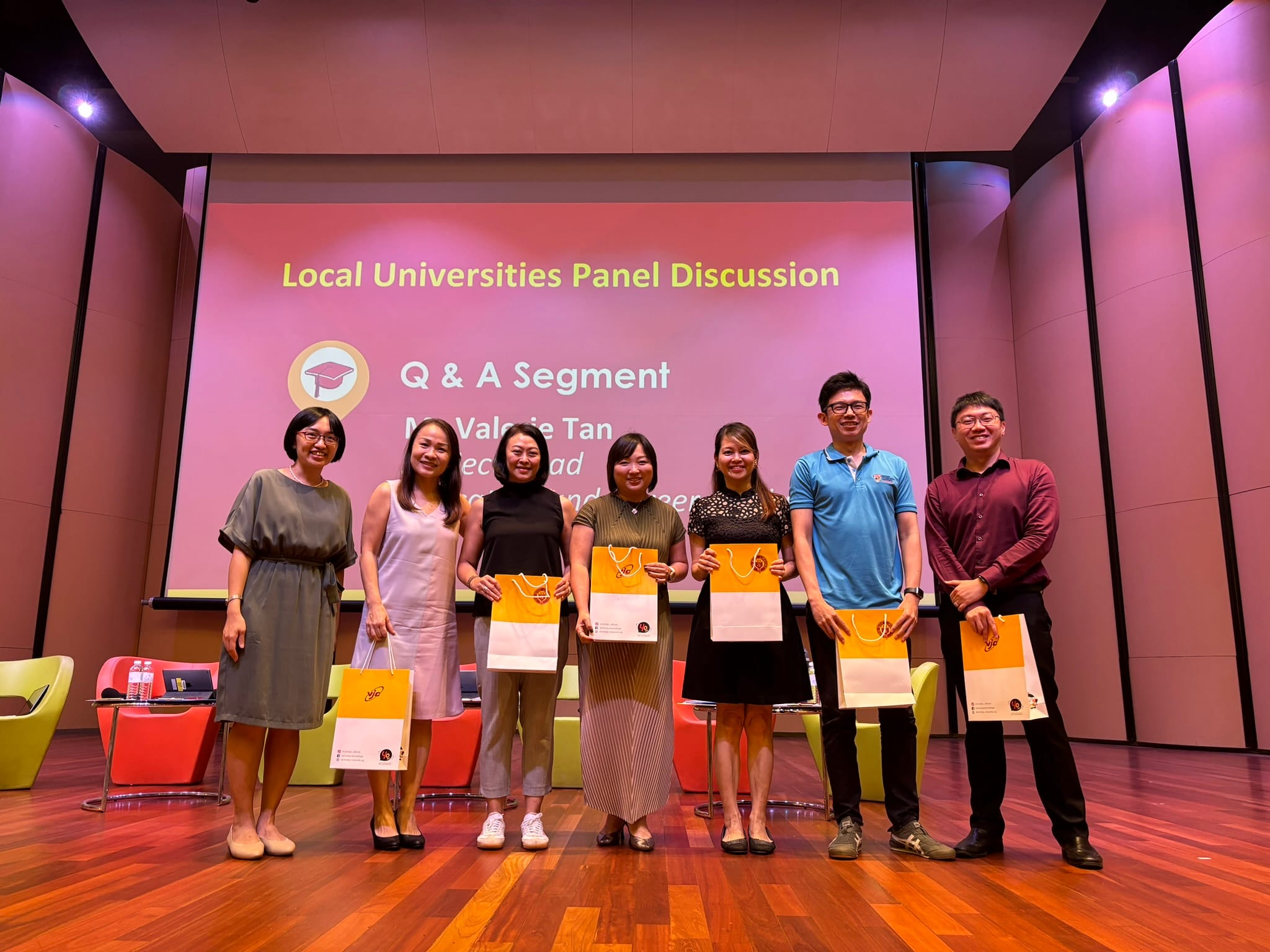 Seven people stand on a stage holding orange gift bags in front of a Local Universities Panel Discussion Q&A screen.