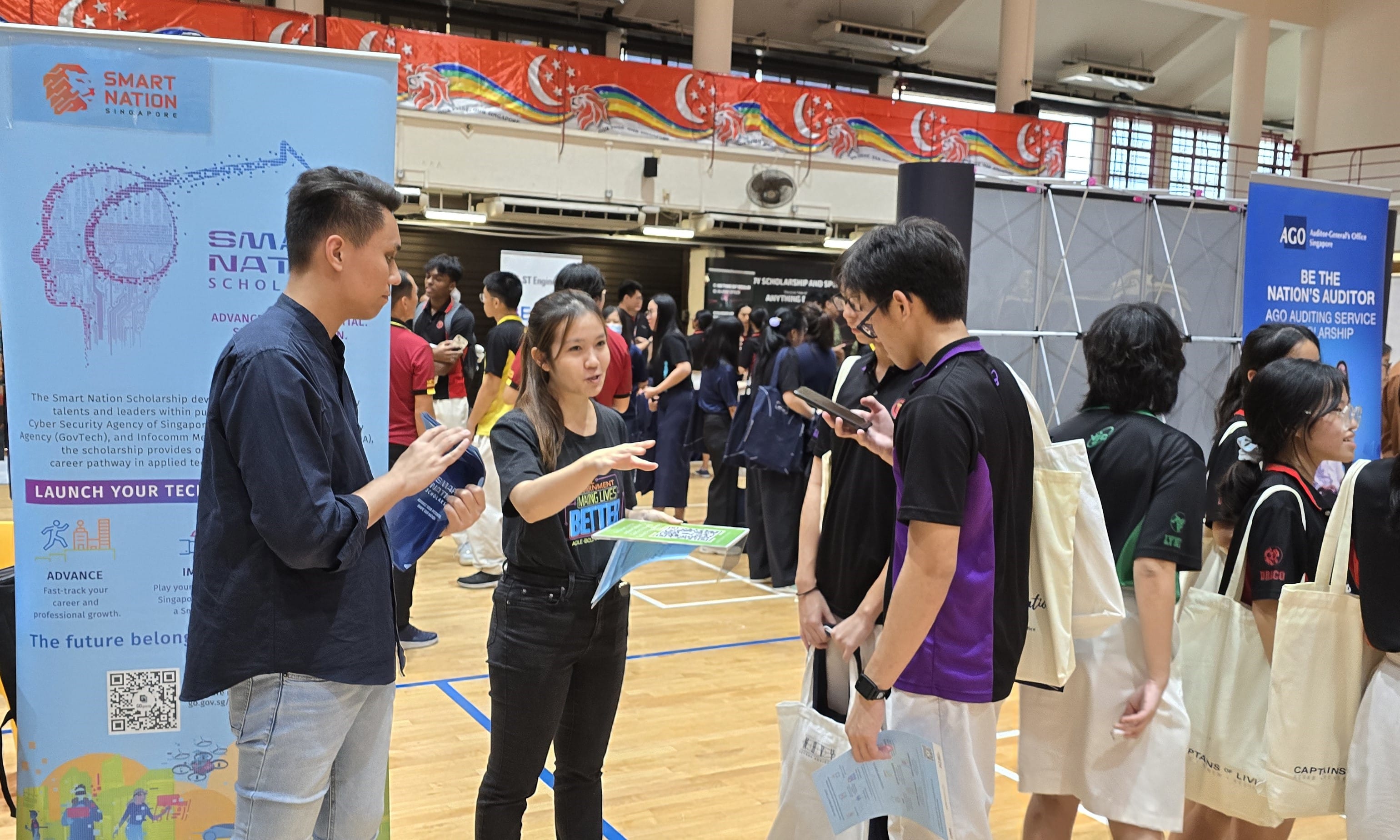 Three young people converse near a Smart Nation Singapore scholarship booth at an indoor career fair.