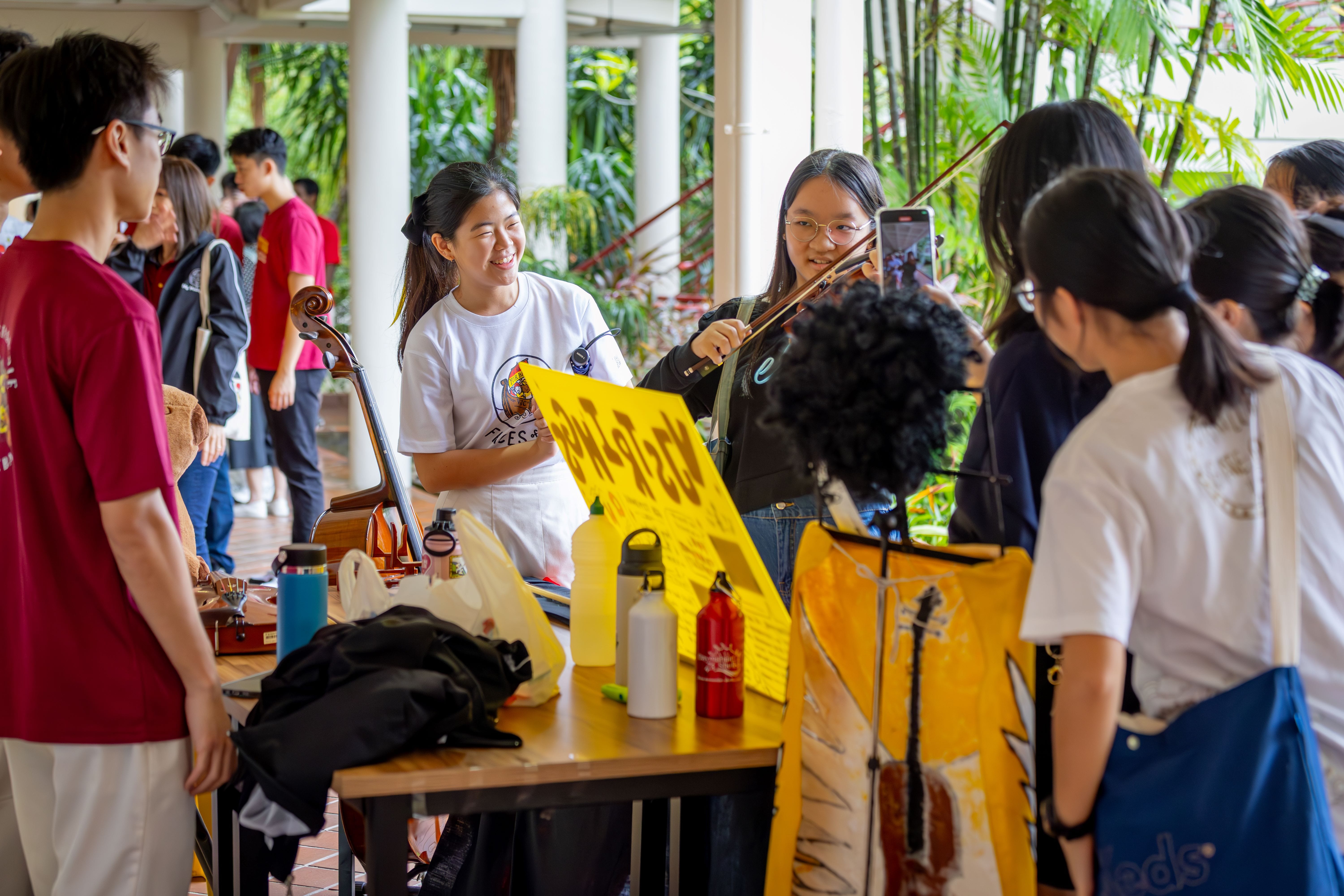 Students gather at an outdoor booth where a girl plays violin beside a smiling girl and a yellow sign.