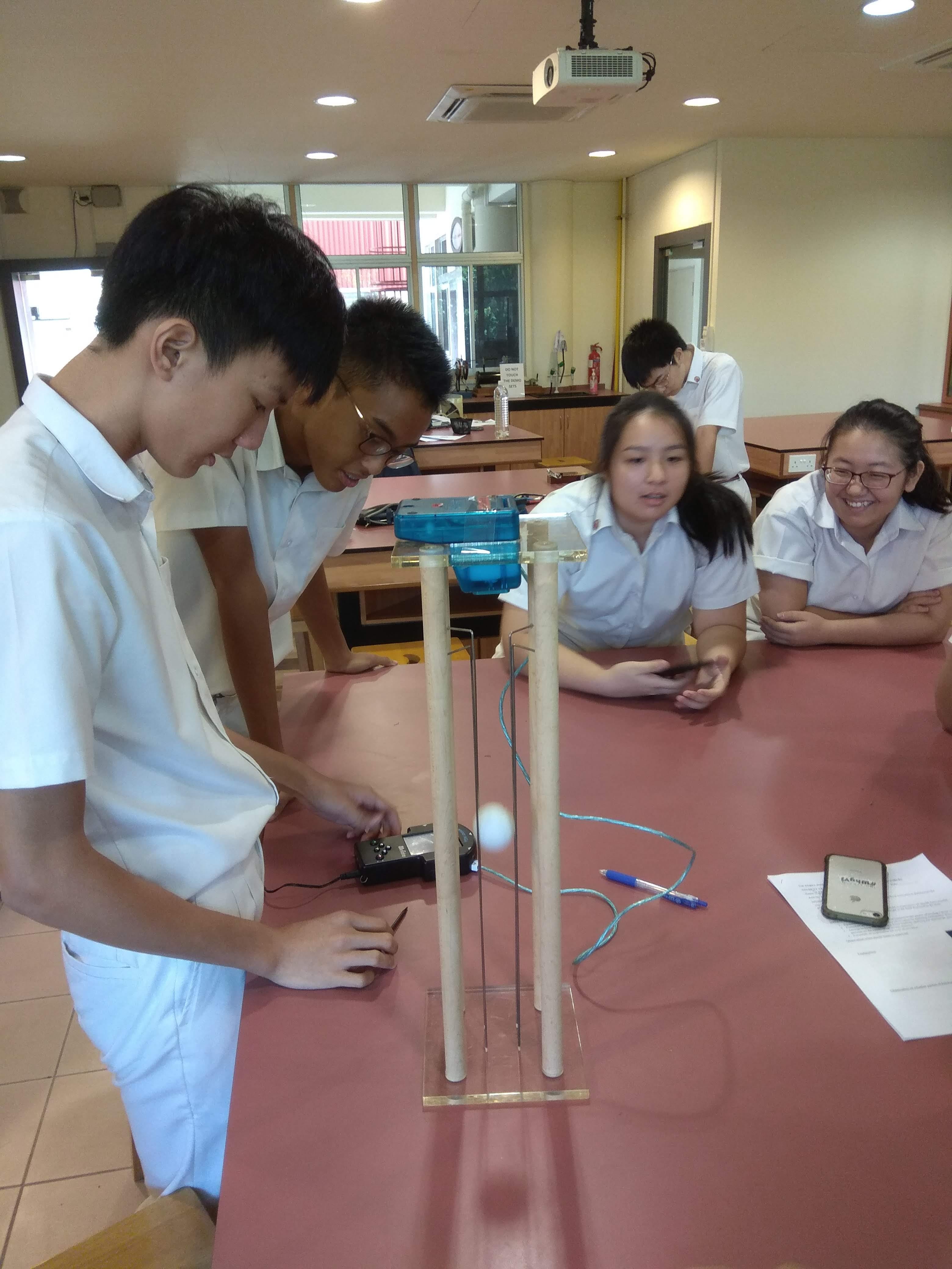 Five students in white uniforms gather around a table conducting a science experiment with a tall wooden stand and a small ball.