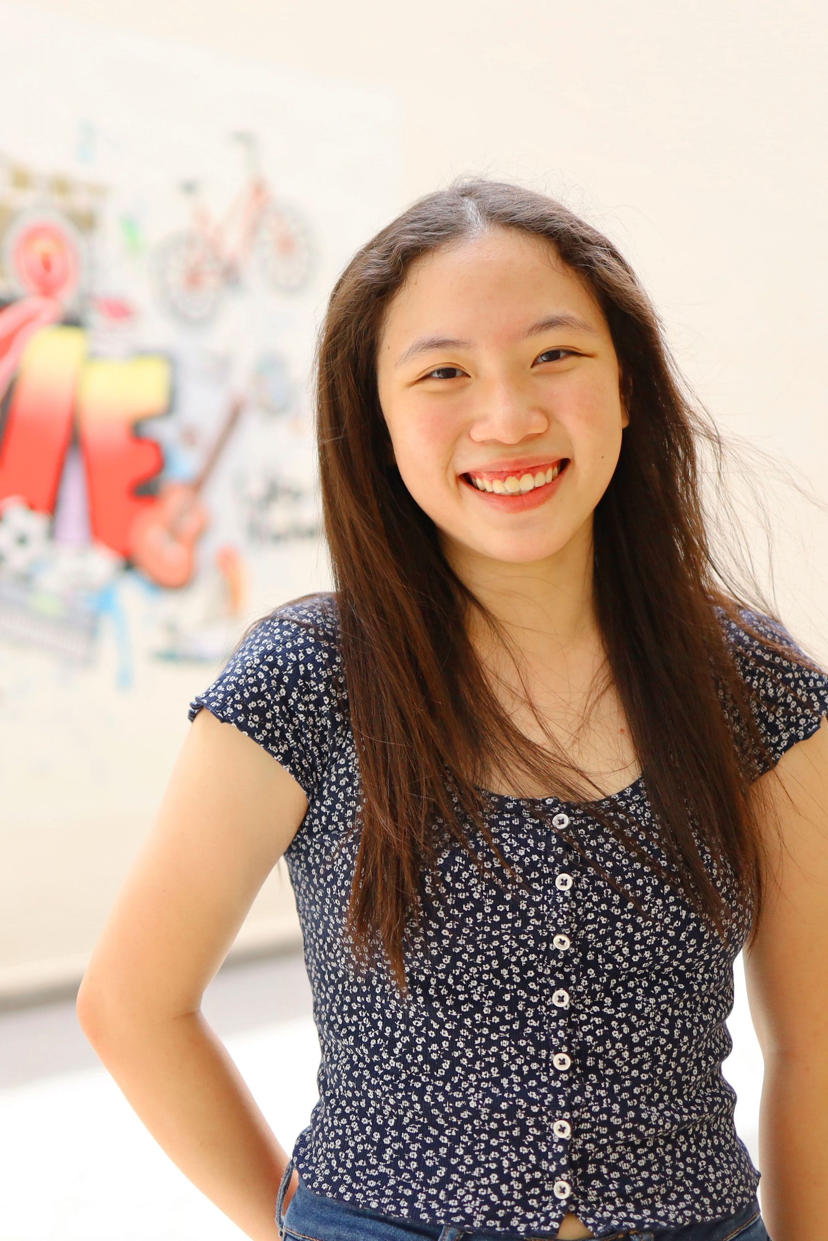 Young woman with long brown hair smiling, wearing a navy floral top, with a blurred colorful mural behind her.