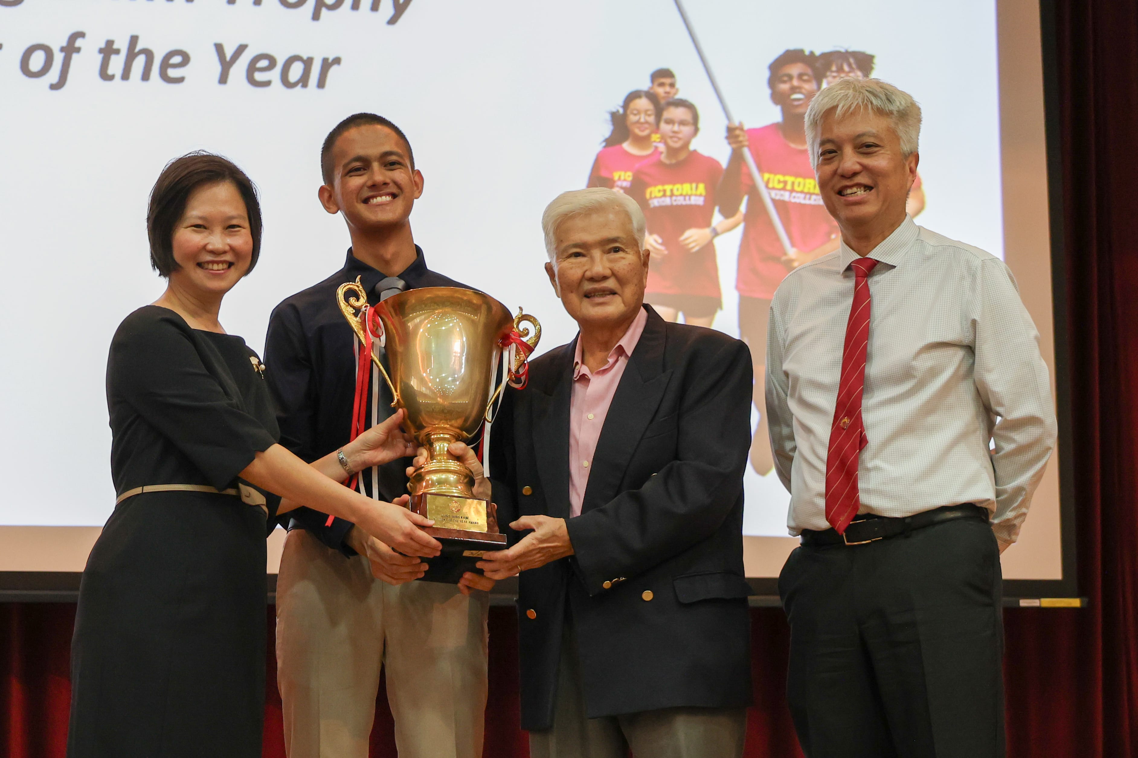 Four people smile on stage as two hold a large gold trophy at a Victoria Junior College award ceremony.