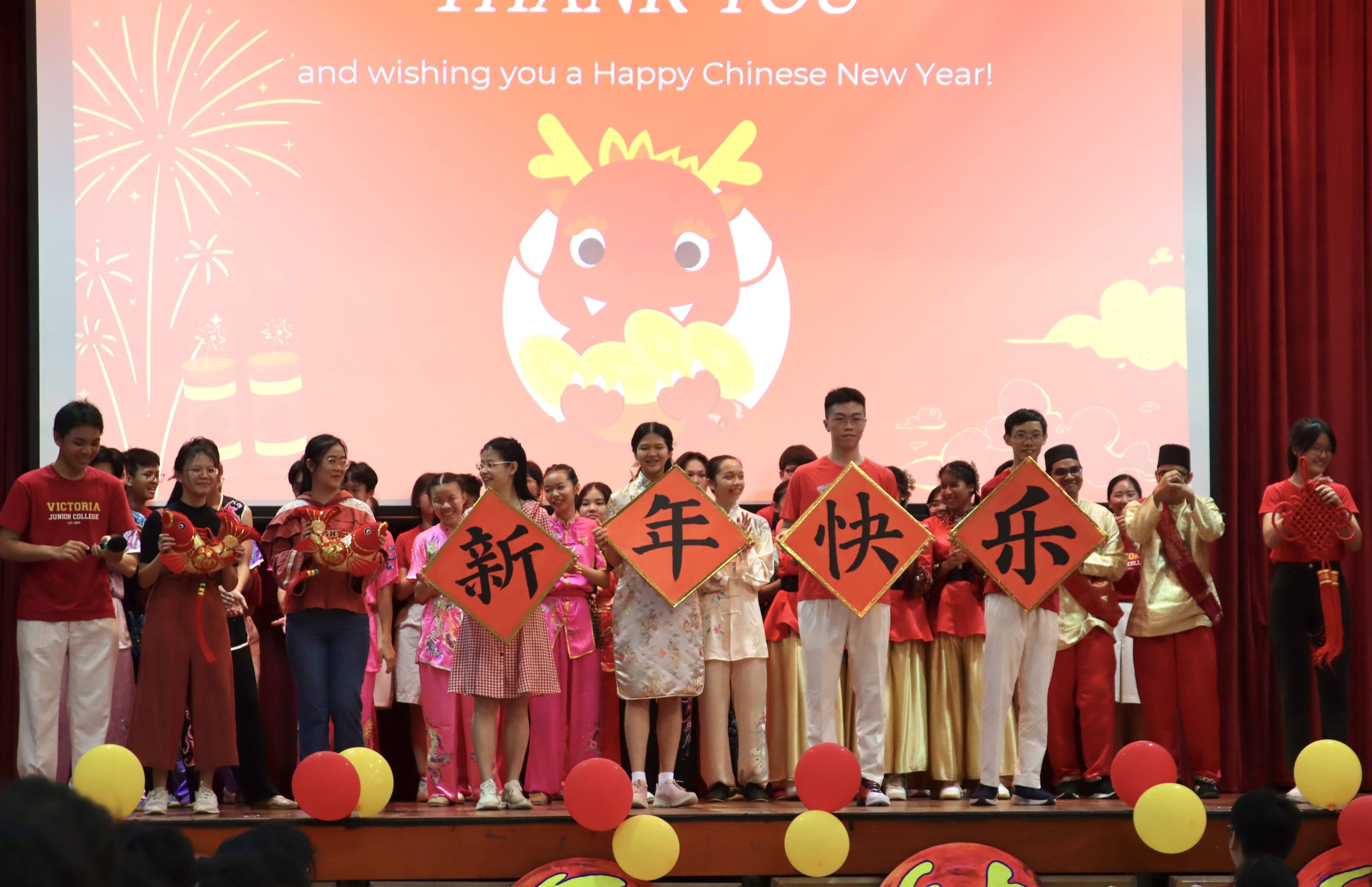 Students on stage hold red signs with Chinese characters meaning Happy New Year at a festive school celebration.