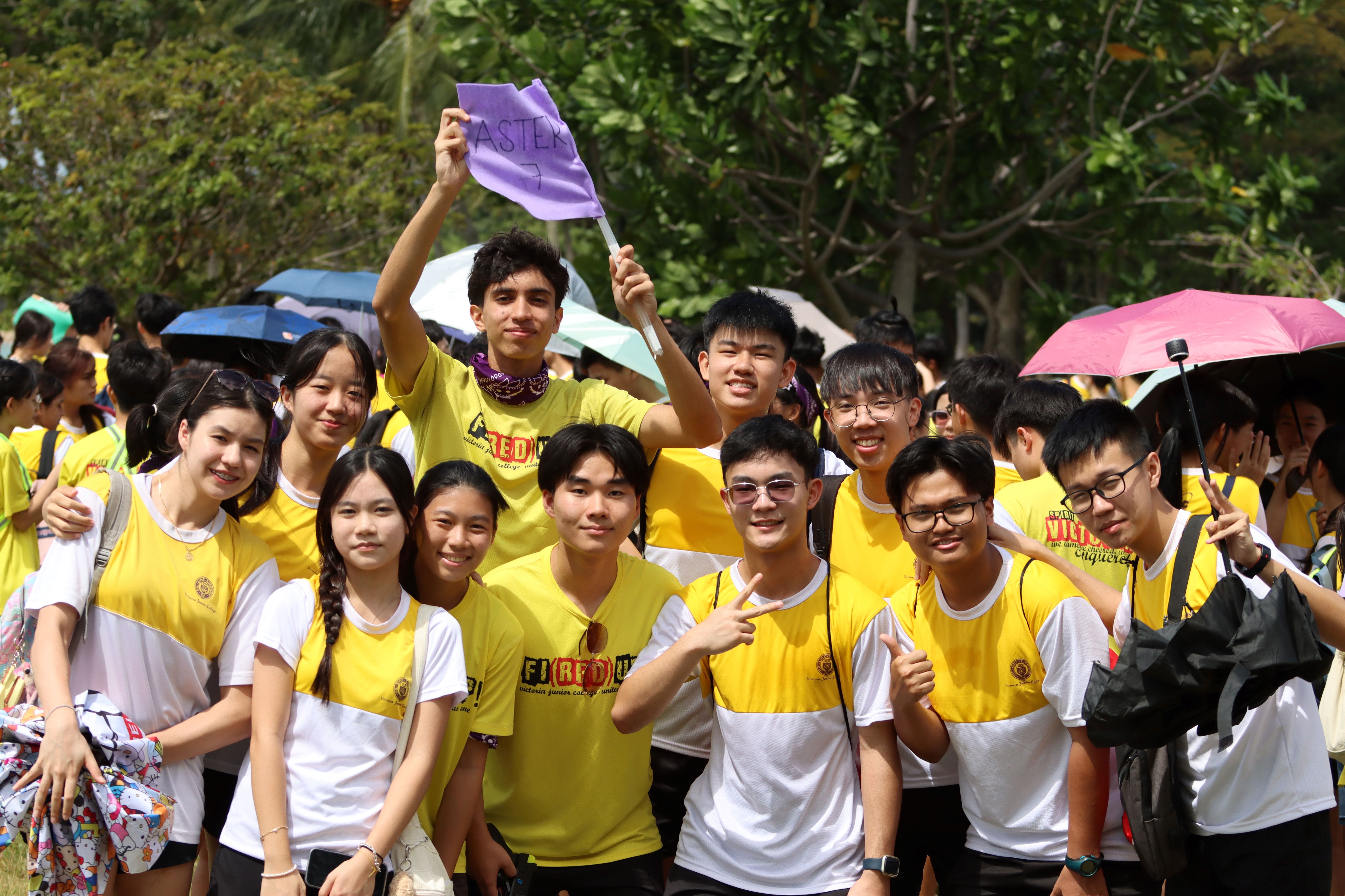 A group of smiling students in yellow and white shirts pose outdoors, one holding a purple flag labeled "Aster 7."