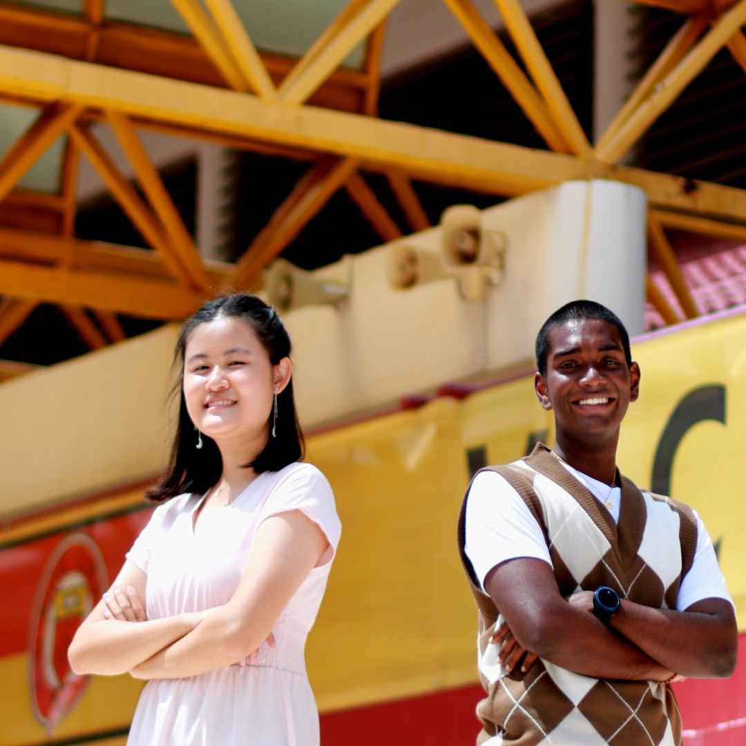 Two smiling young people stand with arms crossed under yellow steel beams near a red and yellow banner.