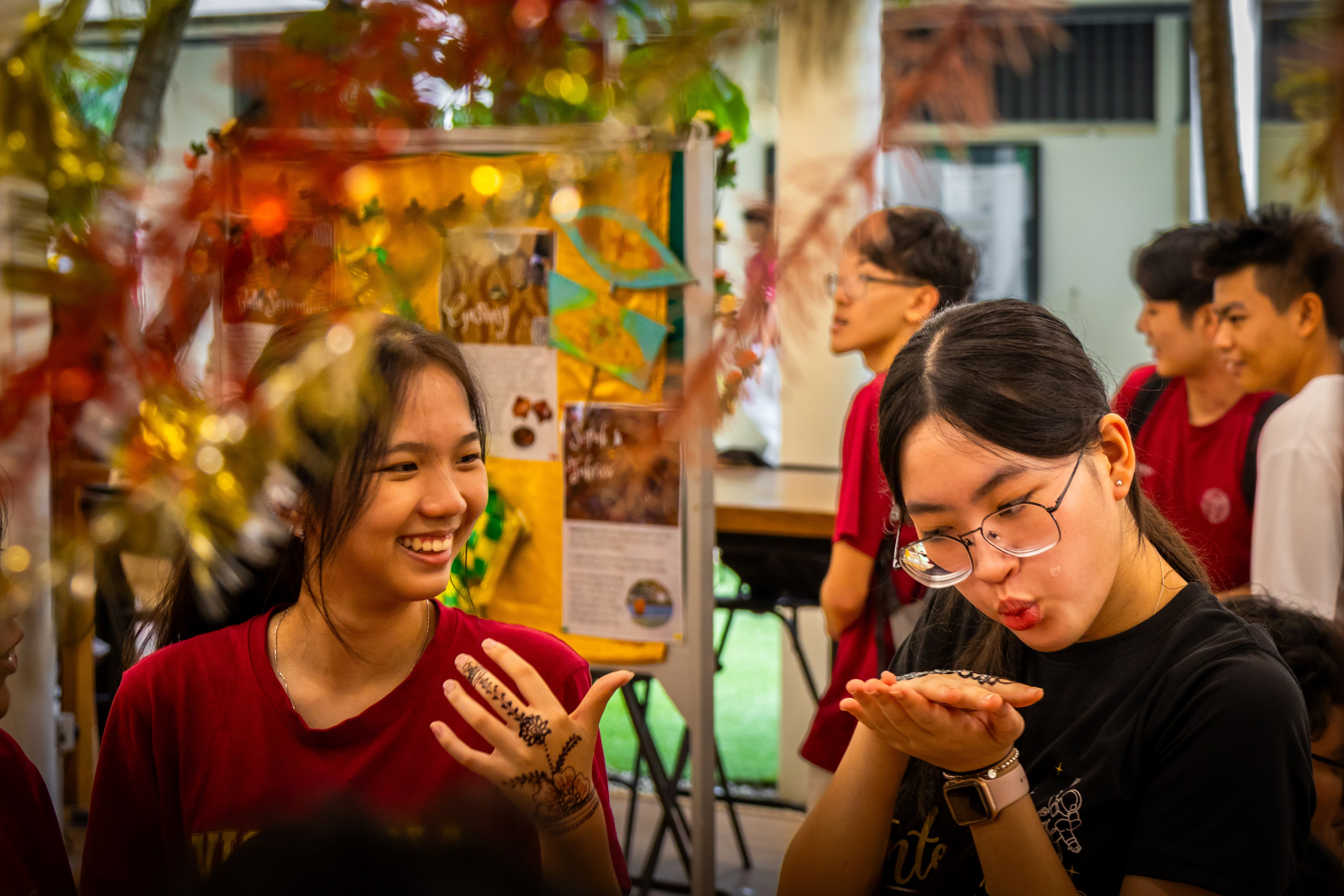Two young women at a festive event, one smiling with henna on her hand, the other blowing something from her palm.