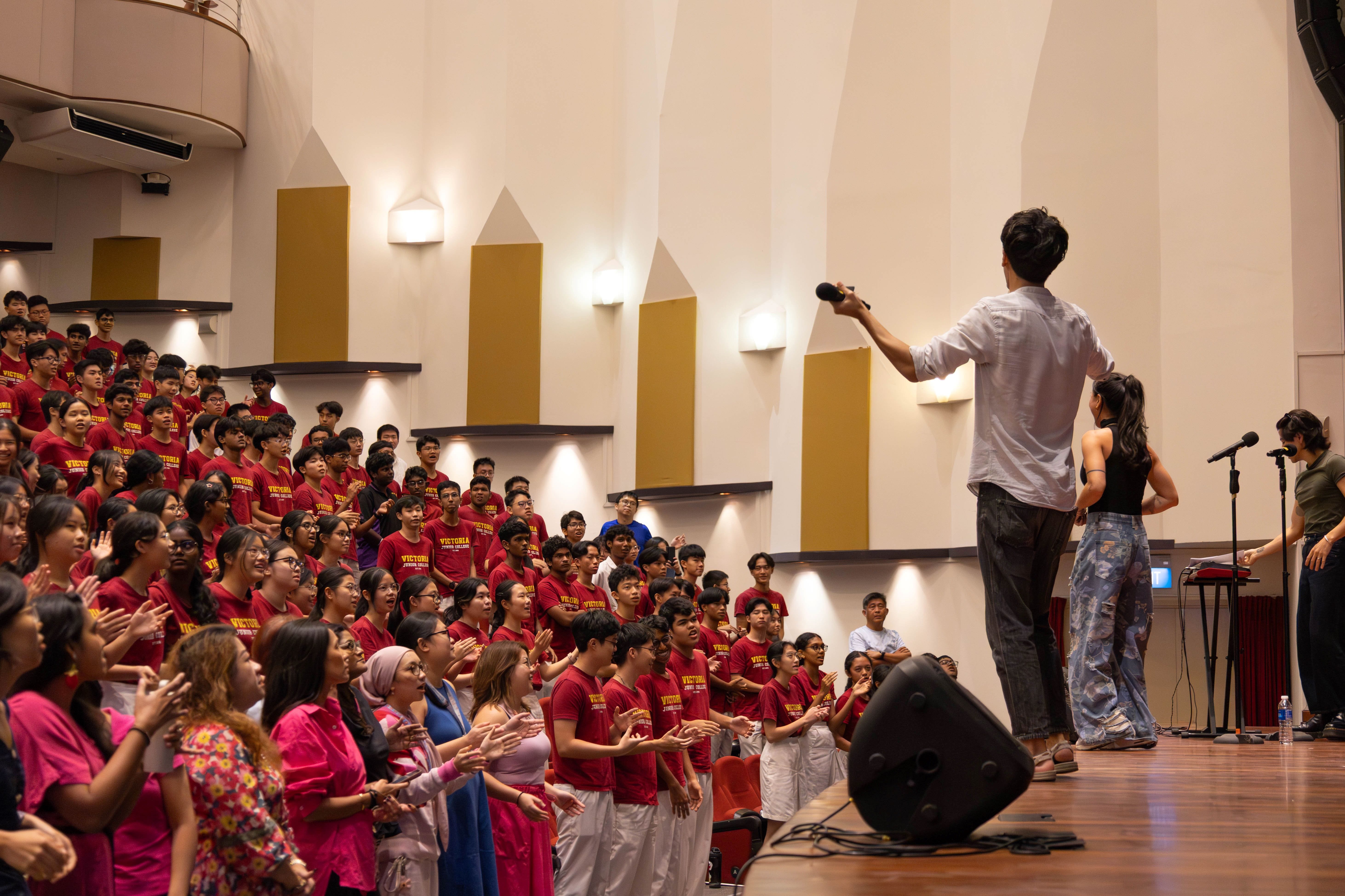A man on stage holds a microphone toward a large crowd of students in red shirts in an auditorium.