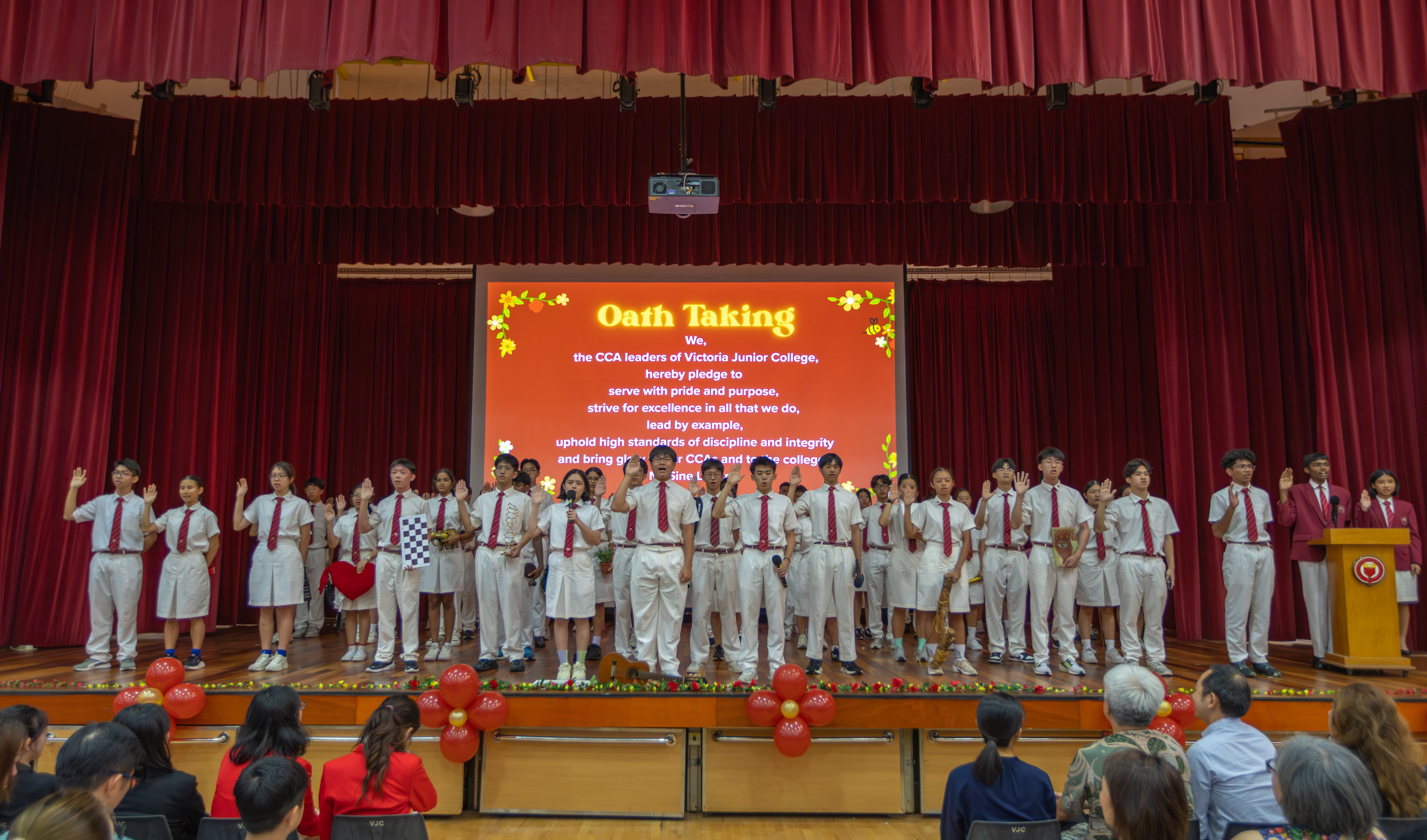 Students in white uniforms raise their right hands on a stage during an Oath Taking ceremony at Victoria Junior College.