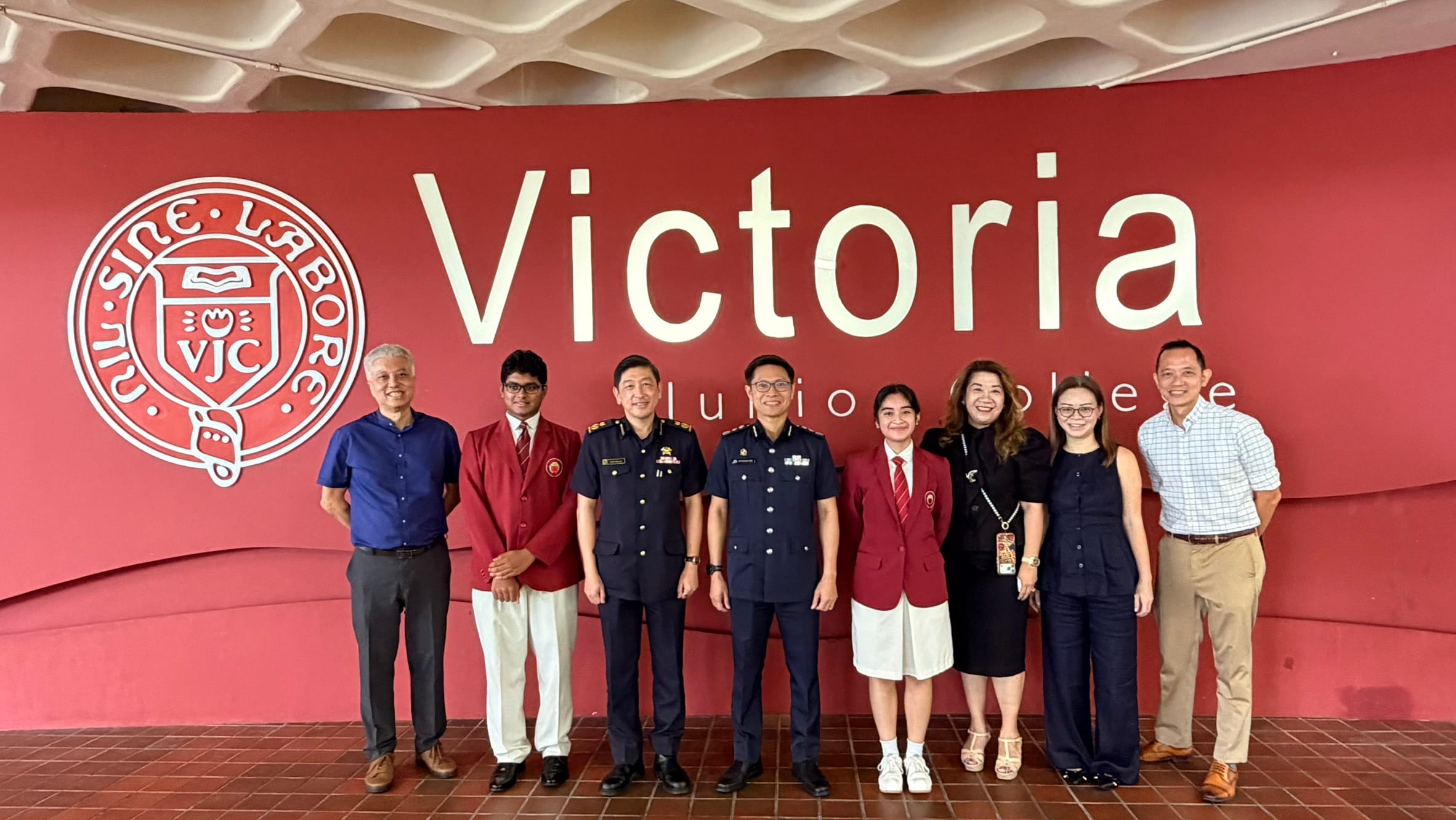 Eight people pose in front of a red Victoria Junior College wall, including two in navy uniforms and two in school blazers.