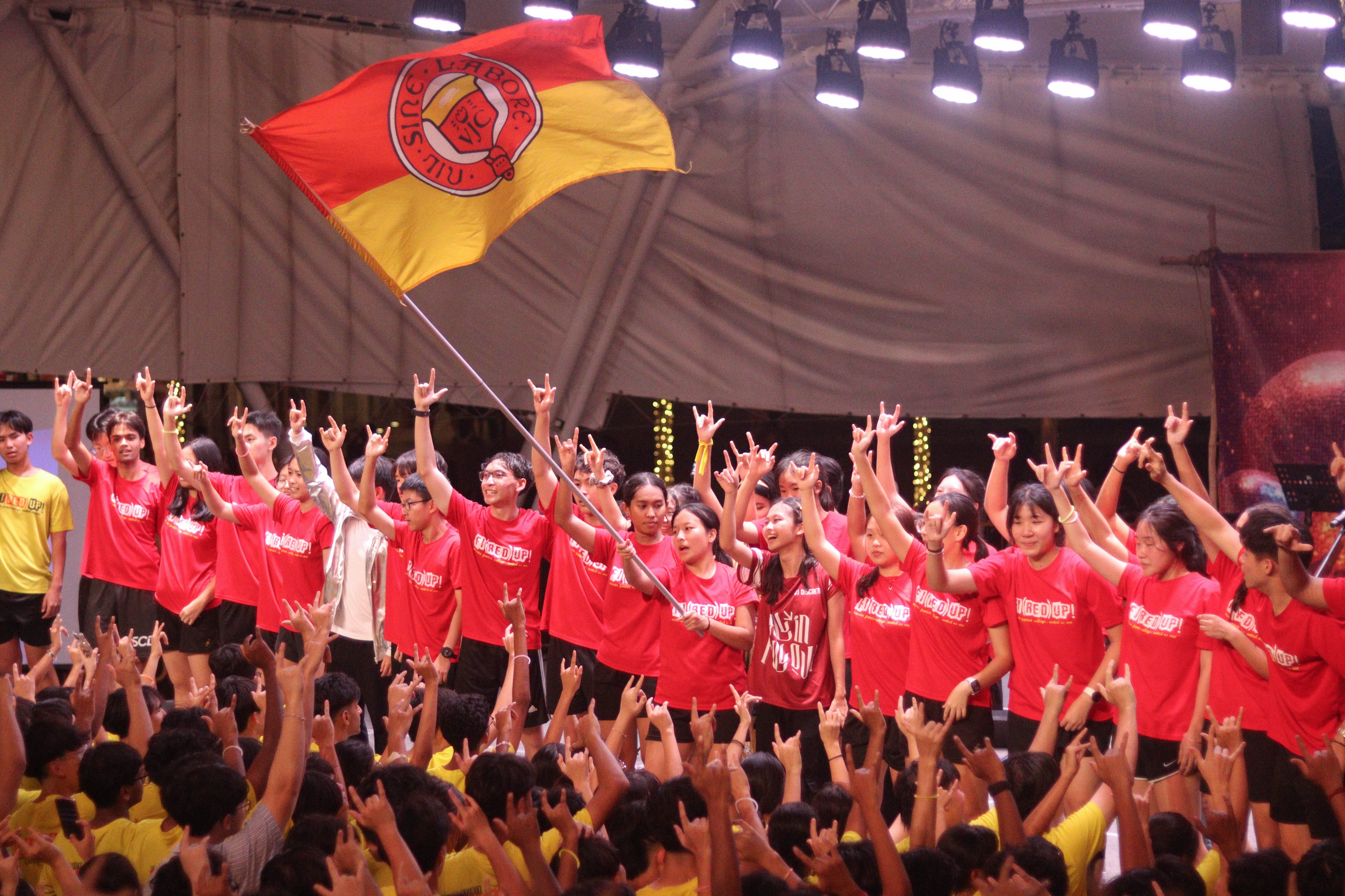 A large group of students in red shirts cheer and raise hands at an event, with one person waving a red and yellow flag.