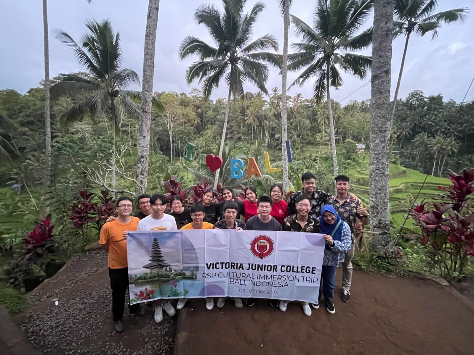A group of students holds a Victoria Junior College Bali cultural trip banner in front of an I Love Bali sign and rice terraces.